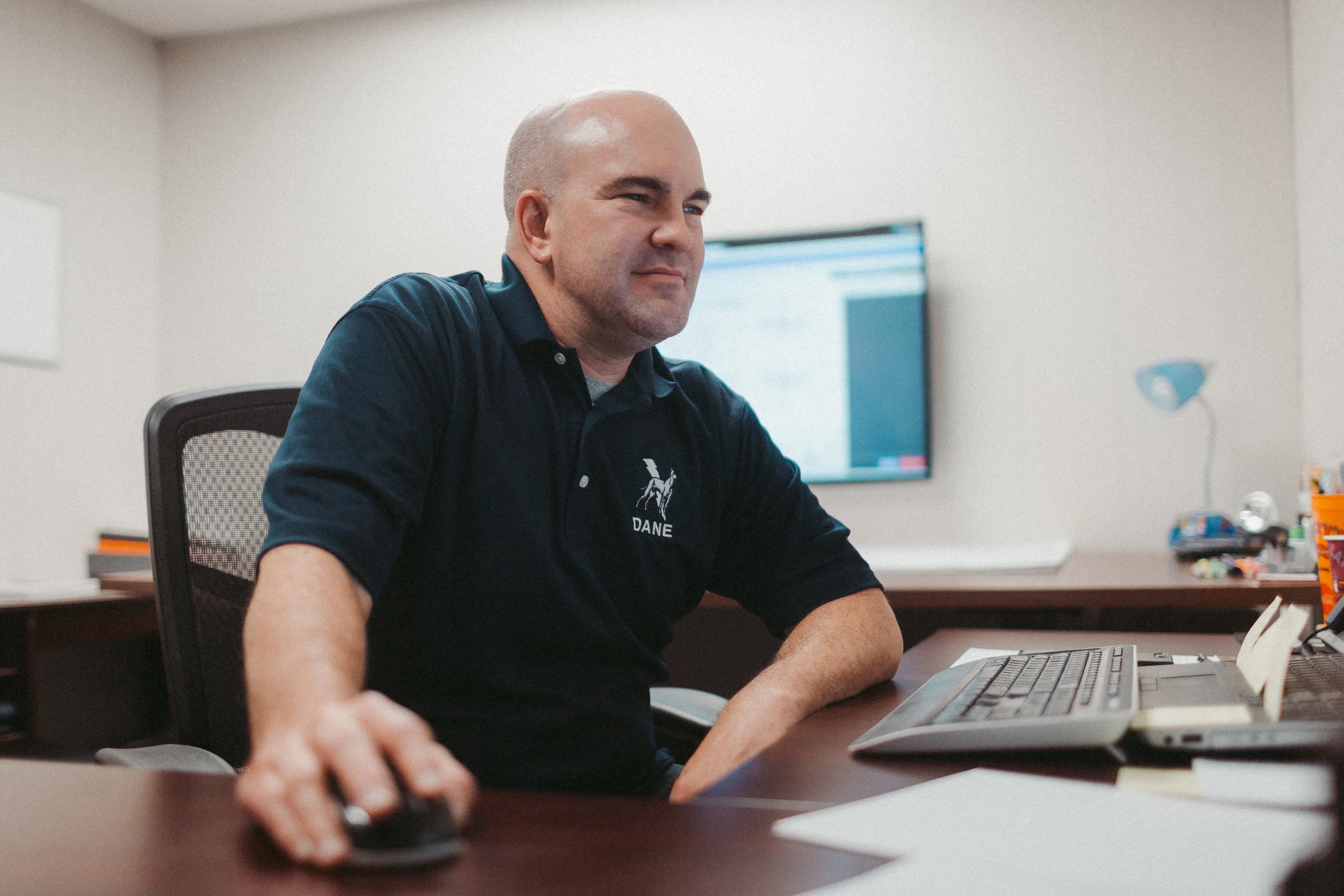 A man with a bald head and light skin sitting at a desk using a computer mouse in an office. He is wearing a navy blue polo shirt with a logo and the name "DANE" on it. There is a laptop and papers on the desk, and a large monitor is visible in the background.