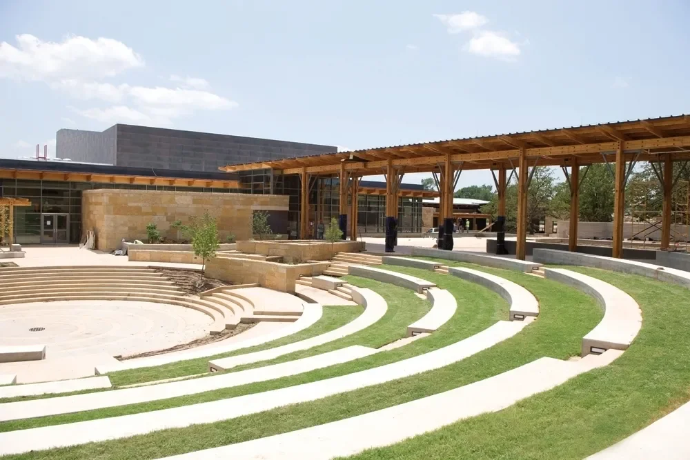 Outdoor amphitheater with curved seating, a grassy area, and a wooden pavilion in the background.