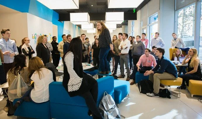 A room full of people standing and sitting on furniture. Shannon Varcoe, a woman with blonde curly hair wearing jeans and a long sleeve shirt stands on a small table to speak
