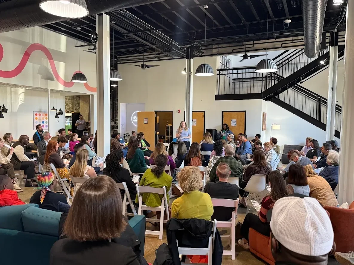 A room full of people in folding chairs. Shannon Varcoe, a blonde woman with curly hair wearing a light purple shirt and jeans, speaks at the front of the room