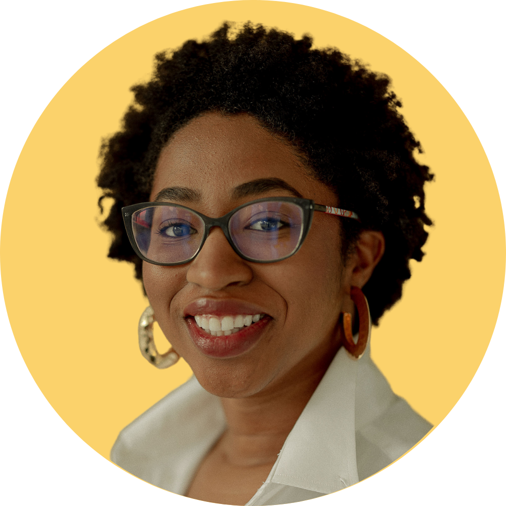 A smiling woman facilitator with dark curly hair, wearing glasses, earrings, and a white shirt, against a yellow circular background. She is one of the three co-founders of women facilitating