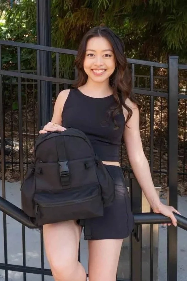 A young woman with long, wavy hair, wearing a sleeveless black top and black shorts, is smiling and holding a black backpack while standing outdoors near a black metal fence with green foliage in the background.