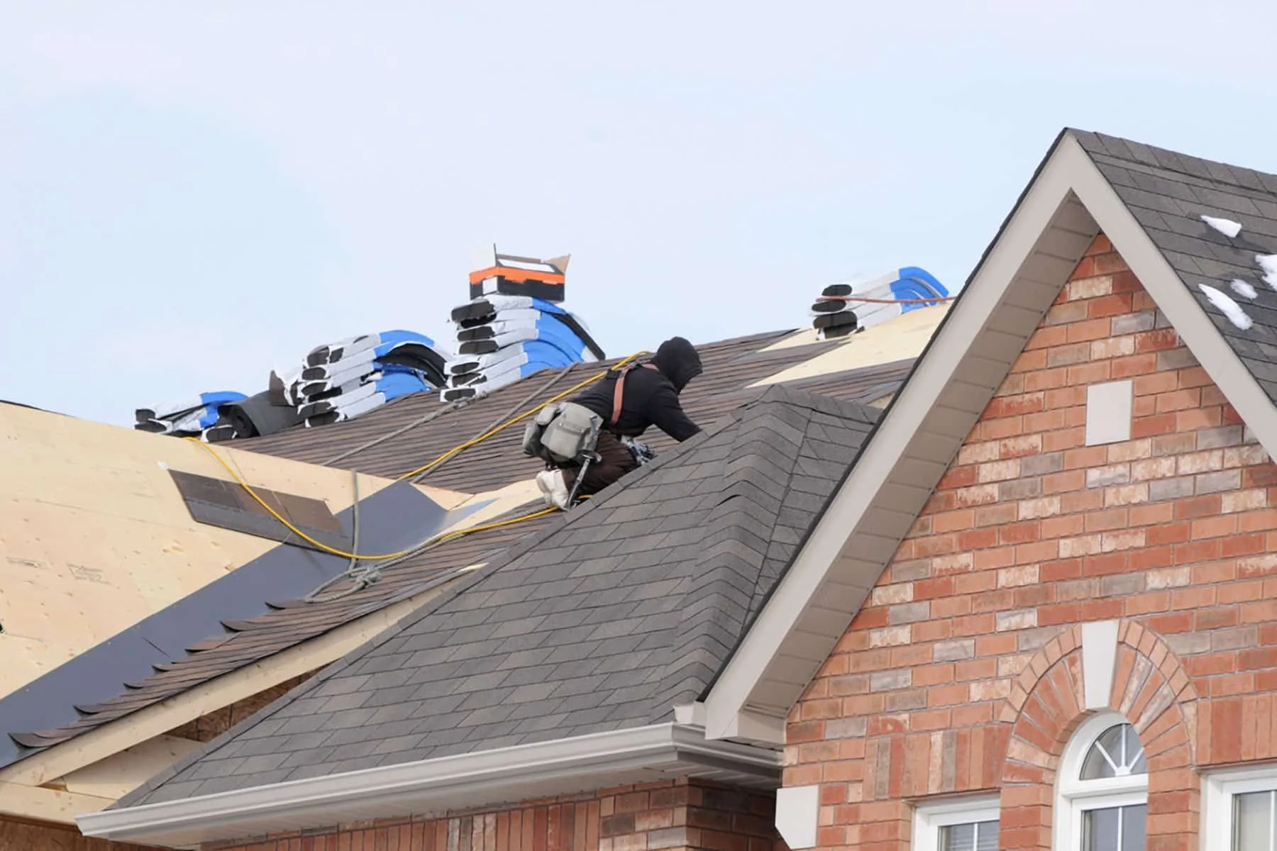 A construction worker wearing a black hoodie and a tool belt is working on the roof of a brick house, installing or repairing roofing shingles. There are stacks of shingles and construction materials on the roof, with roofing tools and materials in t