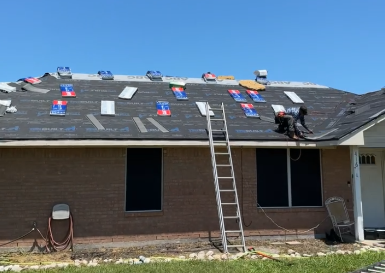 Two workers installing new roofing on a house, with shingles and tools on the roof, ladder leaning against the house, and a clear blue sky.