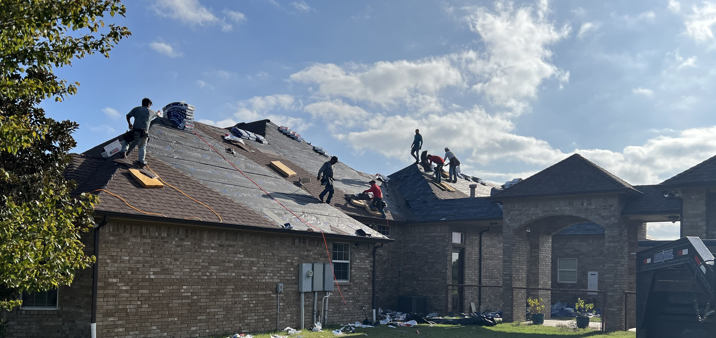 Construction workers installing new roofing on a residential house, with some workers on the roof and others on the ground, under a partly cloudy sky.