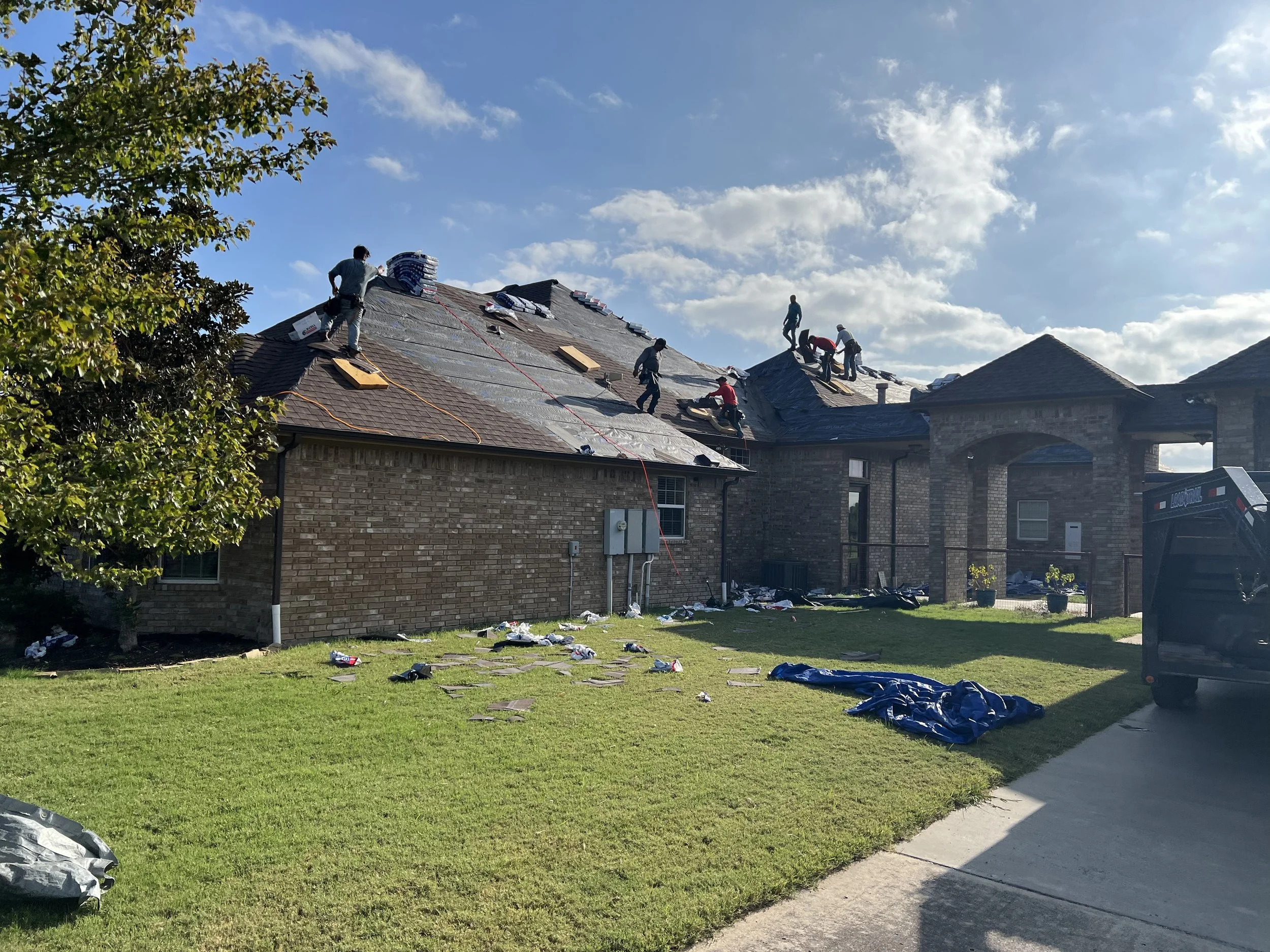People working on the roof of a brick house, installing or repairing shingles, with tools and materials scattered on the roof and yard.