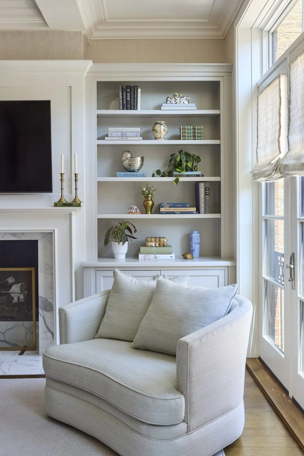 A cozy living room corner featuring a white upholstered armchair, a built-in white bookshelf with decorative items and books, a fireplace with a marble surround, and a large window with light-colored blinds letting in natural light.