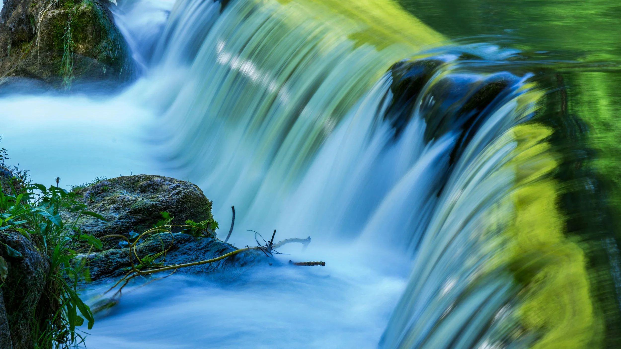 A flowing waterfall over rocks with greenery on the sides, captured with a long exposure to create a smooth water effect.