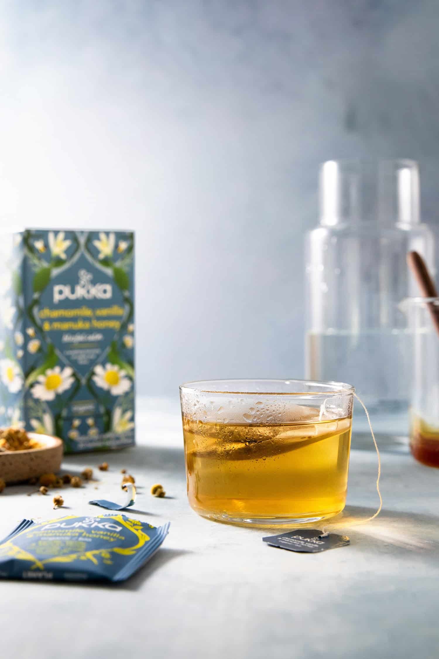 Glass of Pukka chamomile tea with a bottle of water, box of tea, and flower blossoms behind it