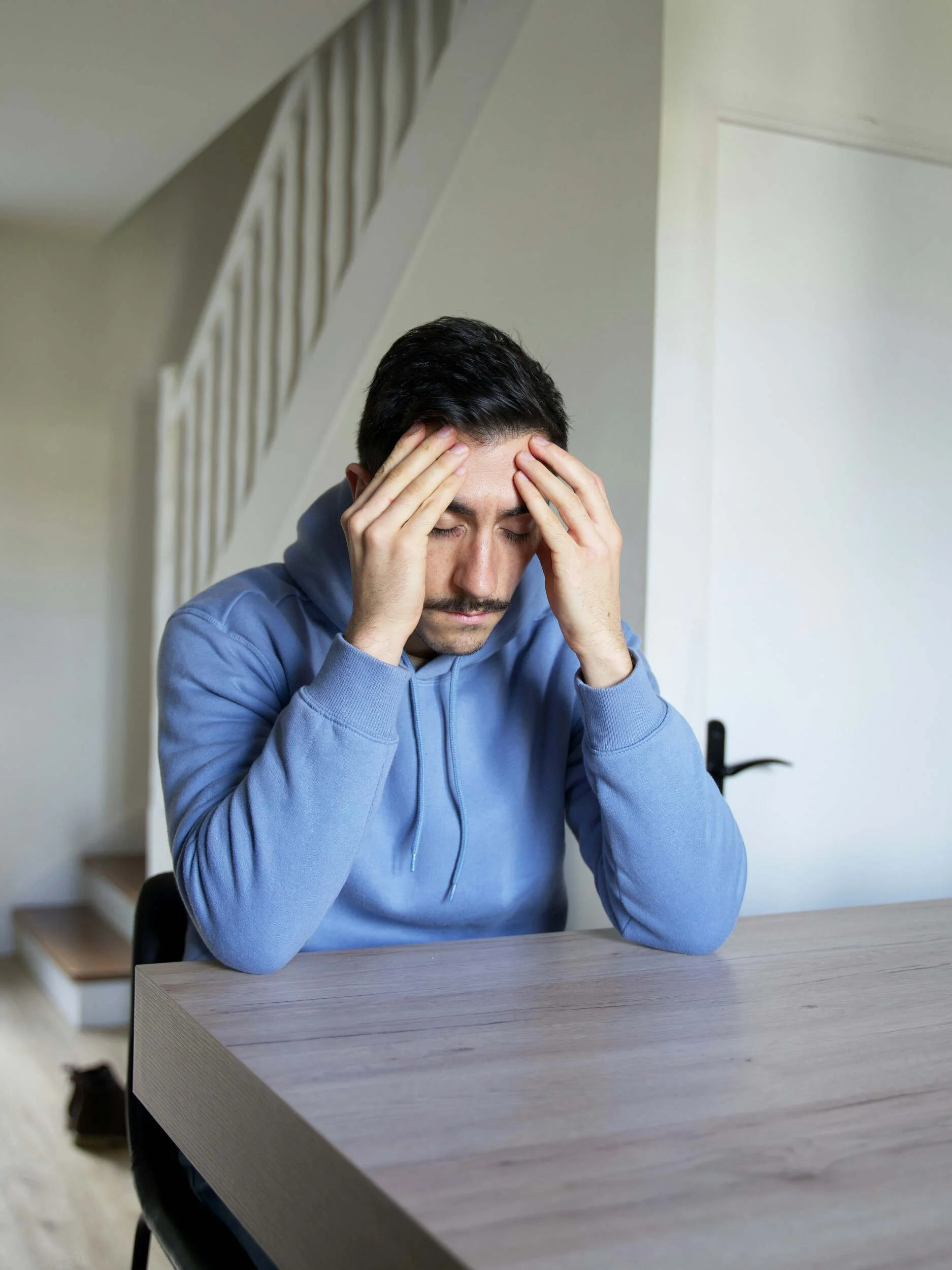 A guy with black hair and a blue sweatshirt sitting at a table inside with his hands on his head - overqualified for a job