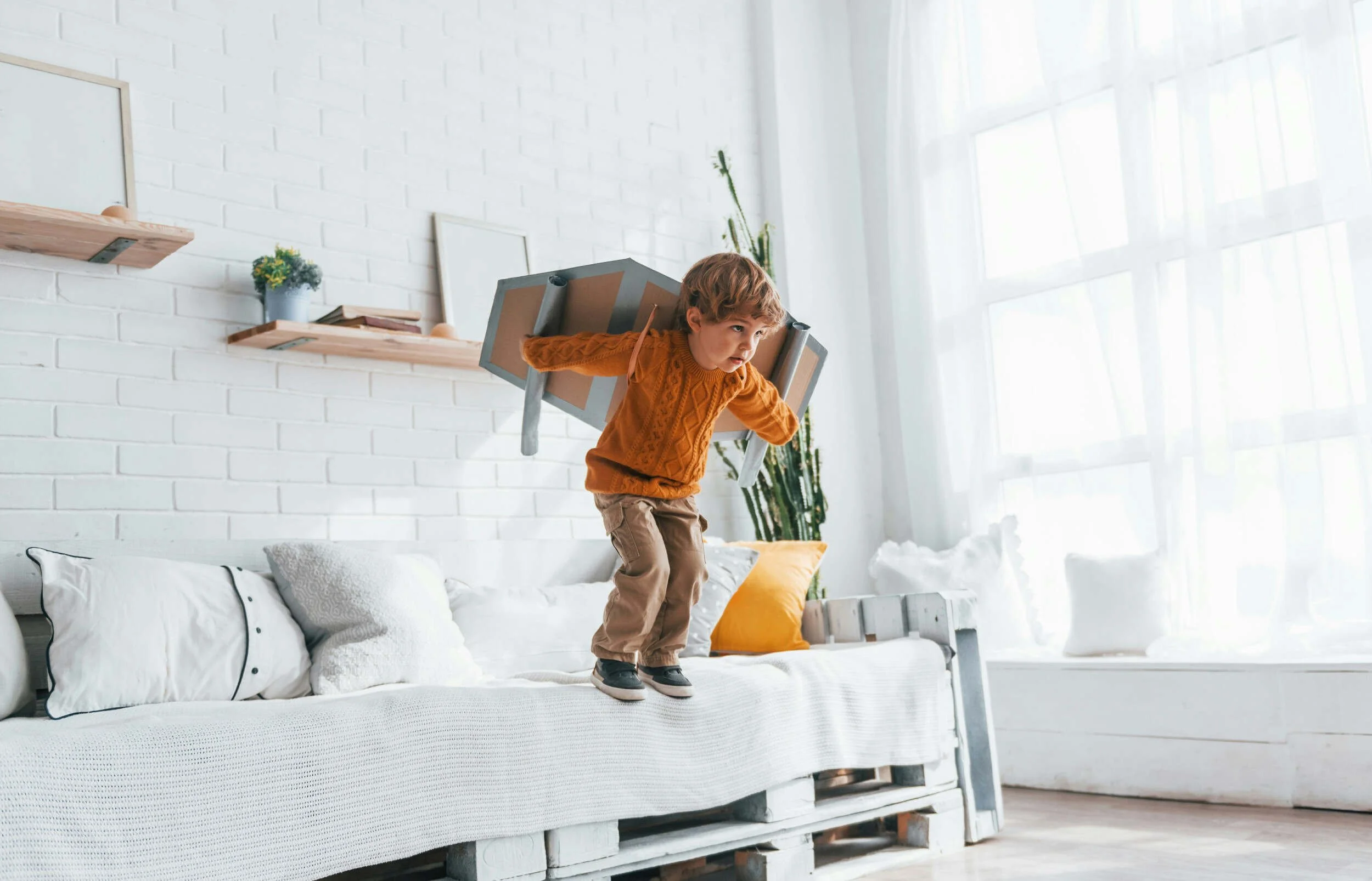 A small child on a couch inside about to jump off with cardboard on his back - successful but unfulfilled