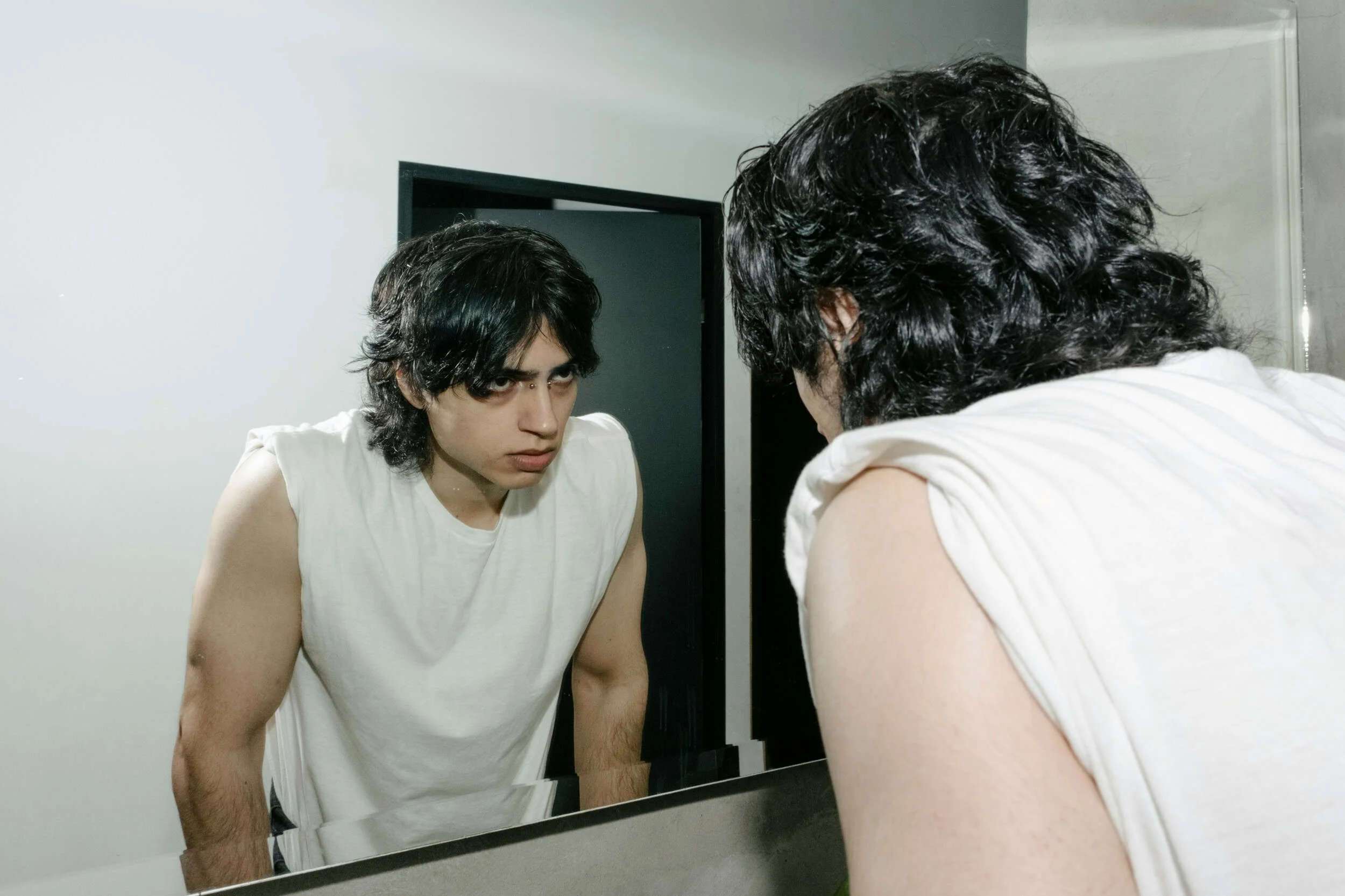 A man with black hair and a white shirt looking in a mirror at himself - How to build a meaningful life