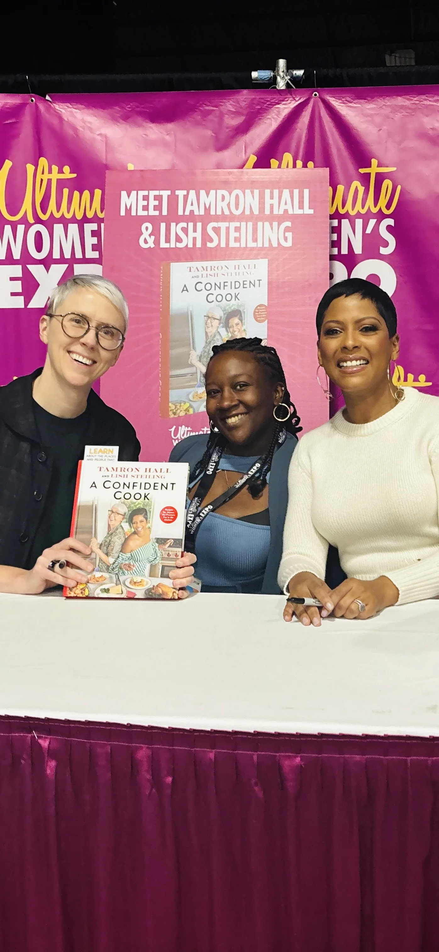 Three women at a book signing for 'A Confident Cook' by Tamron Hall and Lisha Steling, with a pink promotional backdrop and a table covered with a white cloth and a pink curtain.
