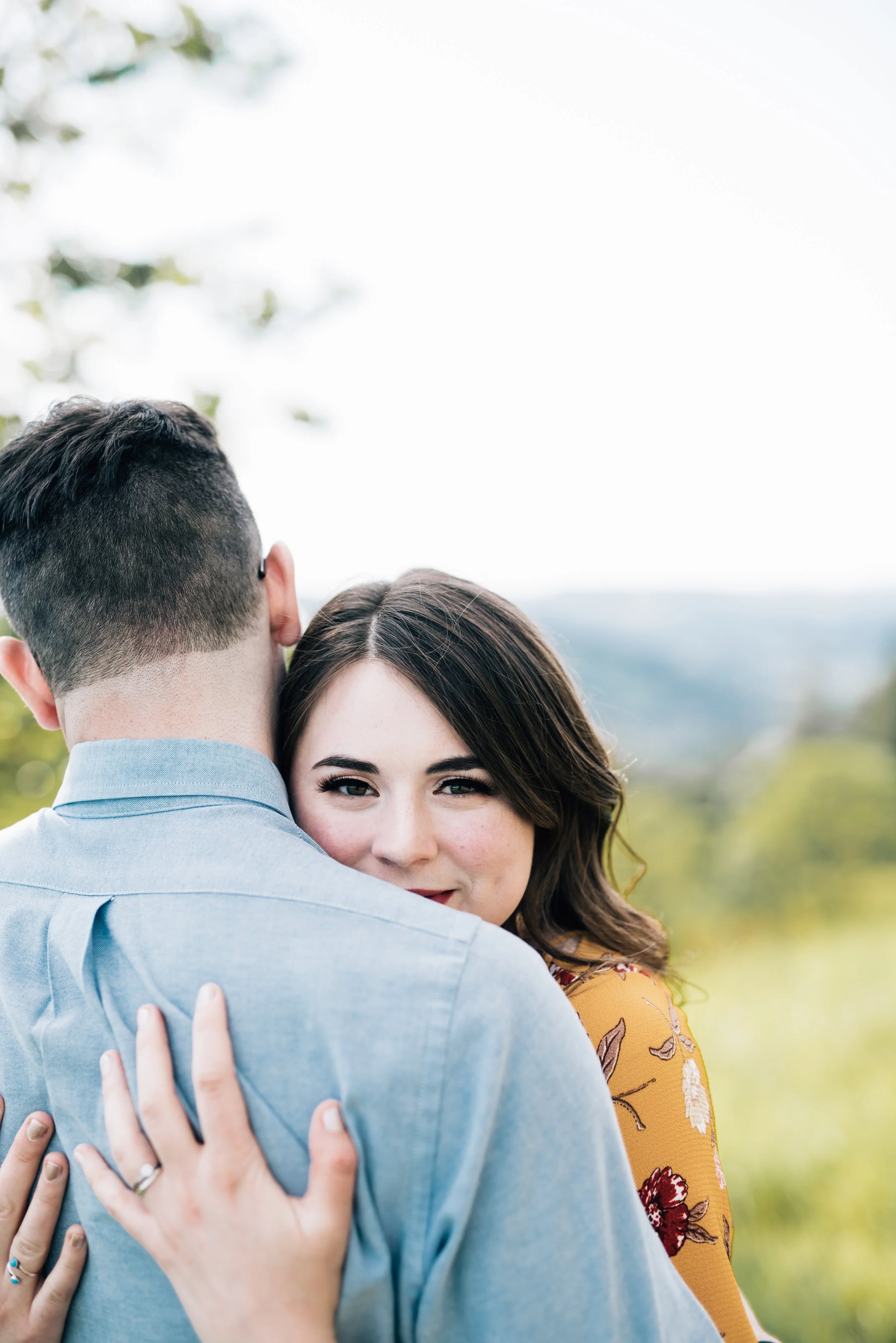 A brunette woman holds onto her husband who is facing the other direction, looking at the camera