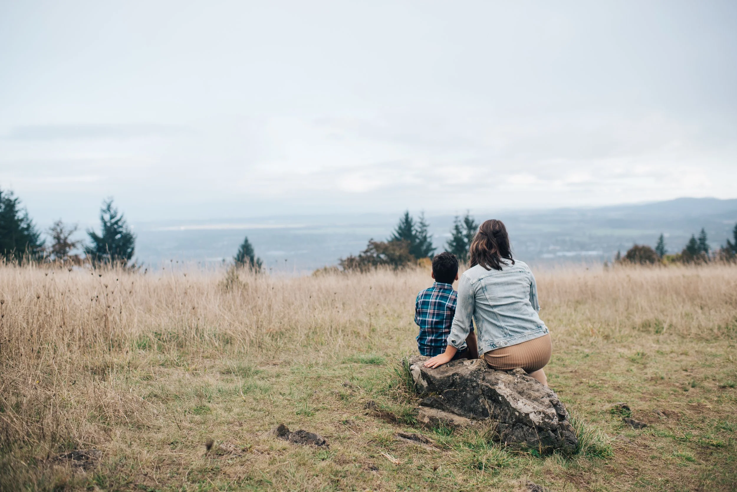 View from behind, a mother and son look off into the distance from atop a hill.