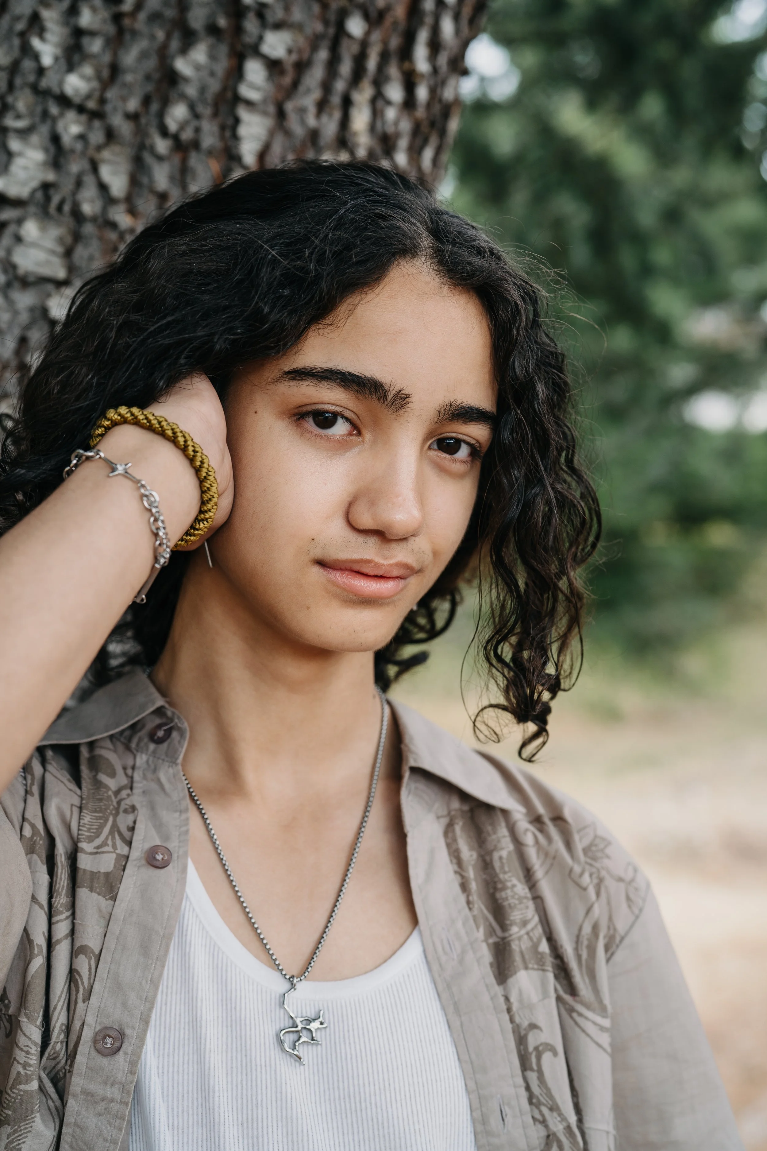 A high school senior leans up against a tree giving a subtle smile while running their hand through their hair.