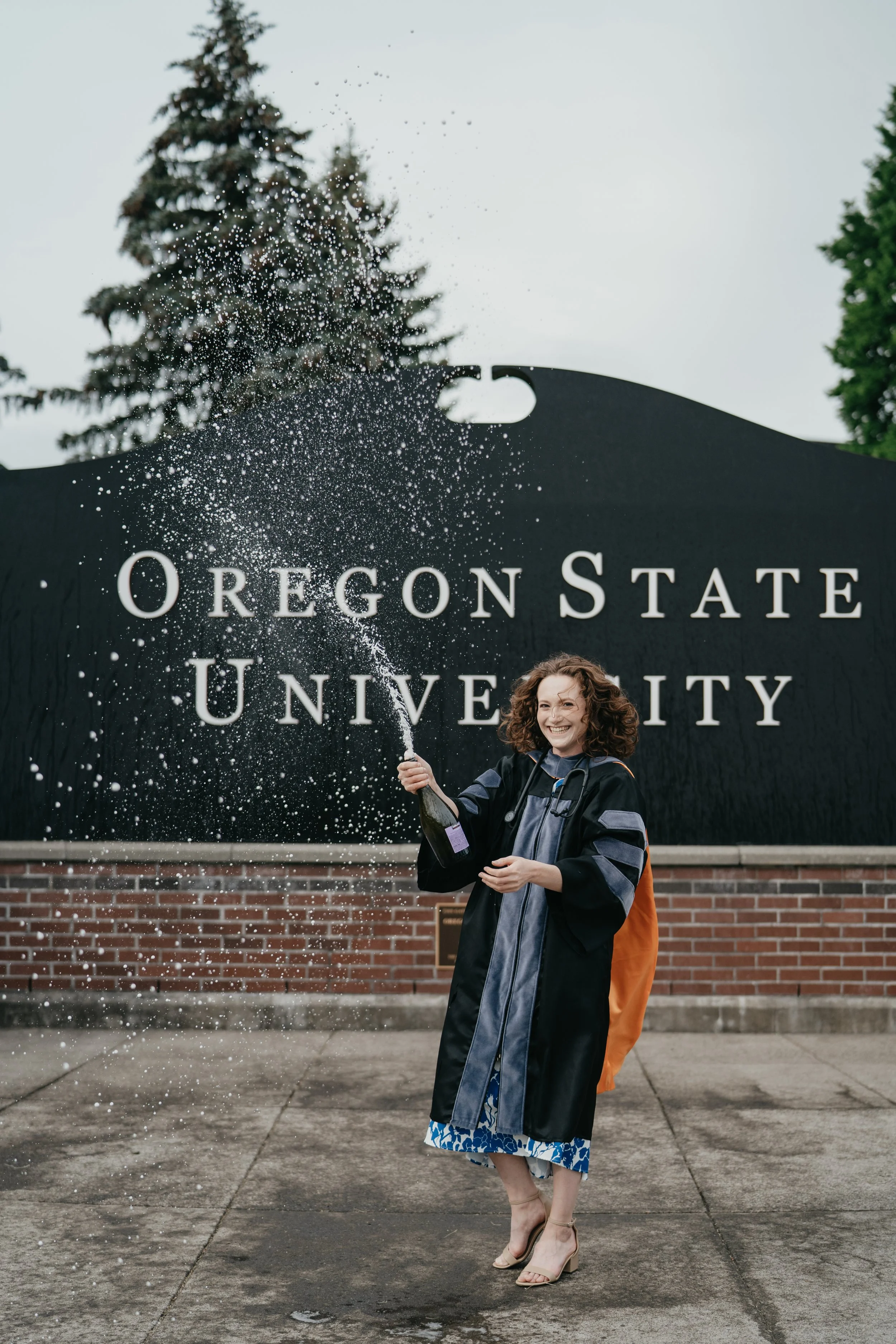A college graduate shoots off a champagne bottle in front of a sign that says Oregon State University