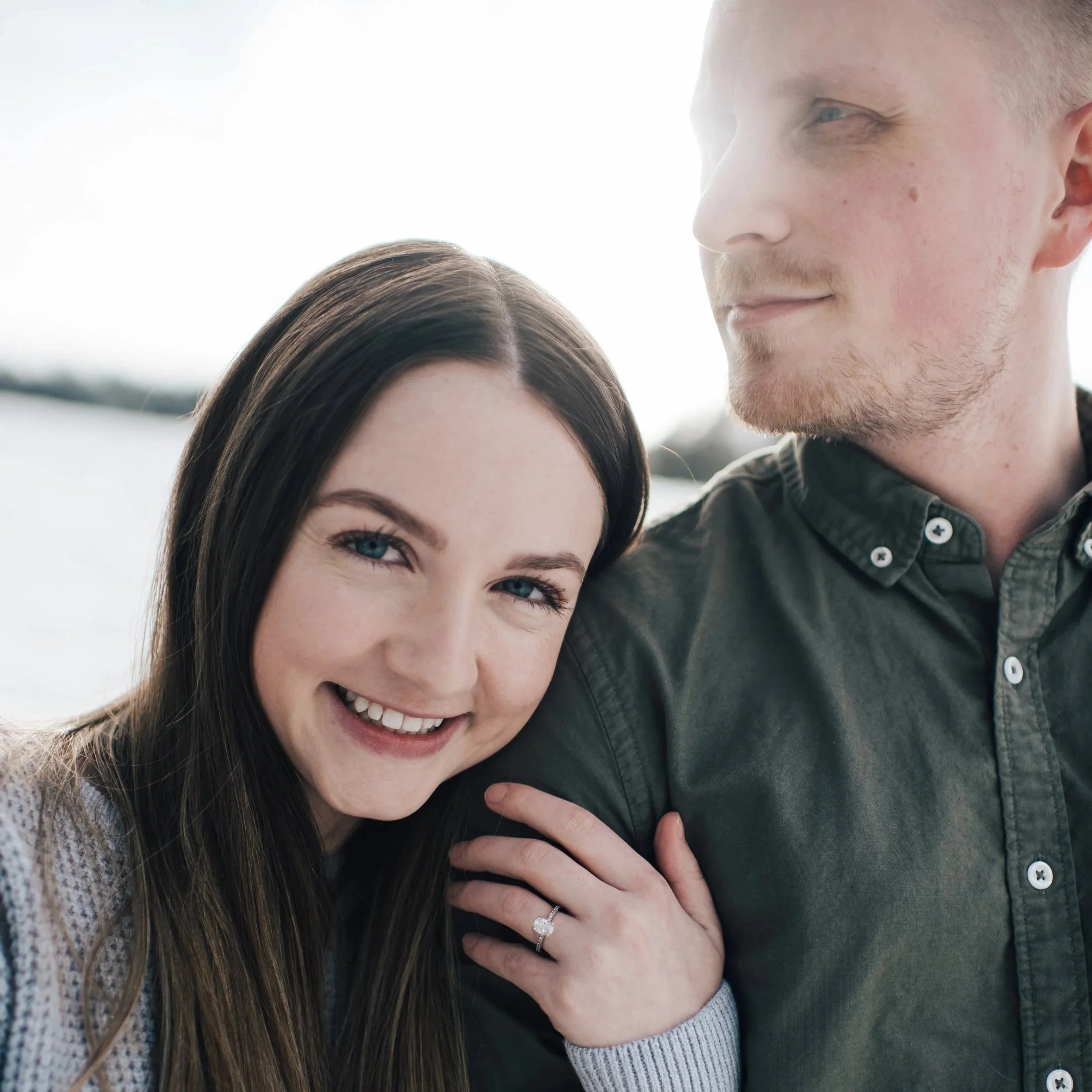 A female with long brown hair has her arm wrapped around her fiance's arm showing off her engagement ring.