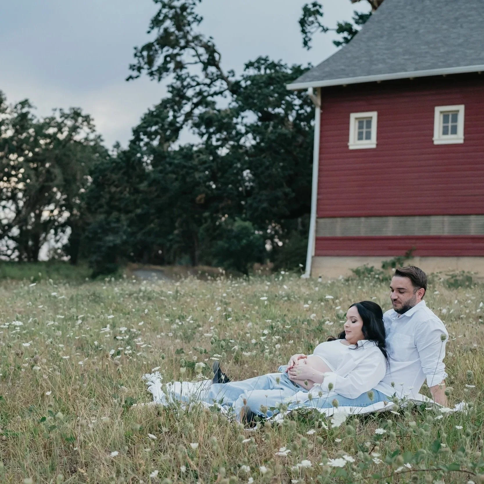 A couple expecting a baby lay in the grass in front of a red barn. 