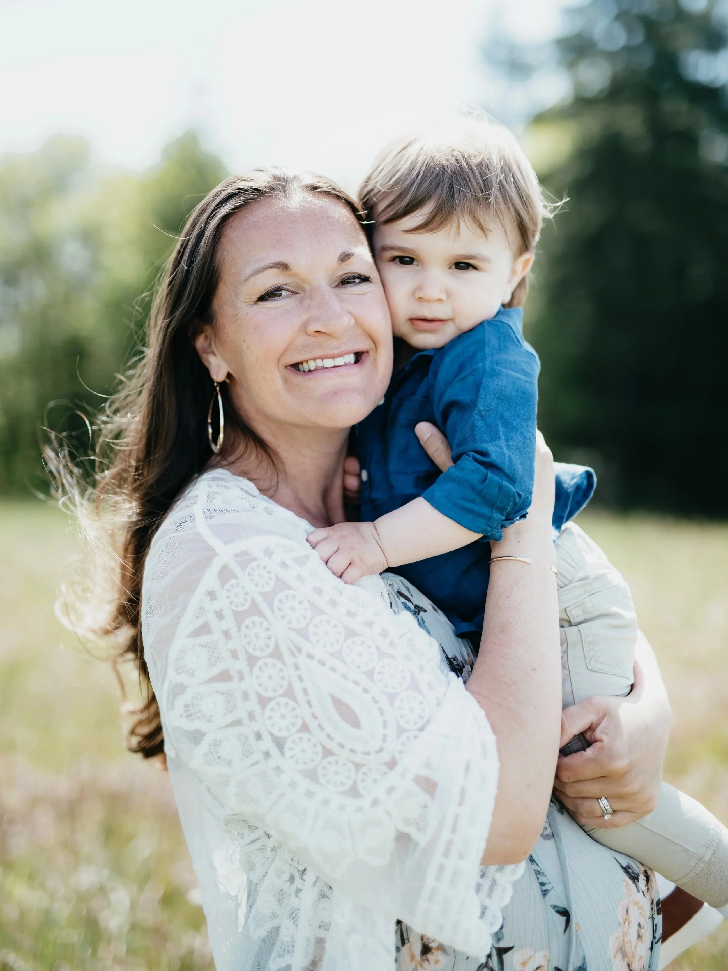 Pregnant woman with long brown hair wearing a white dress smiles while holding her two year old son.