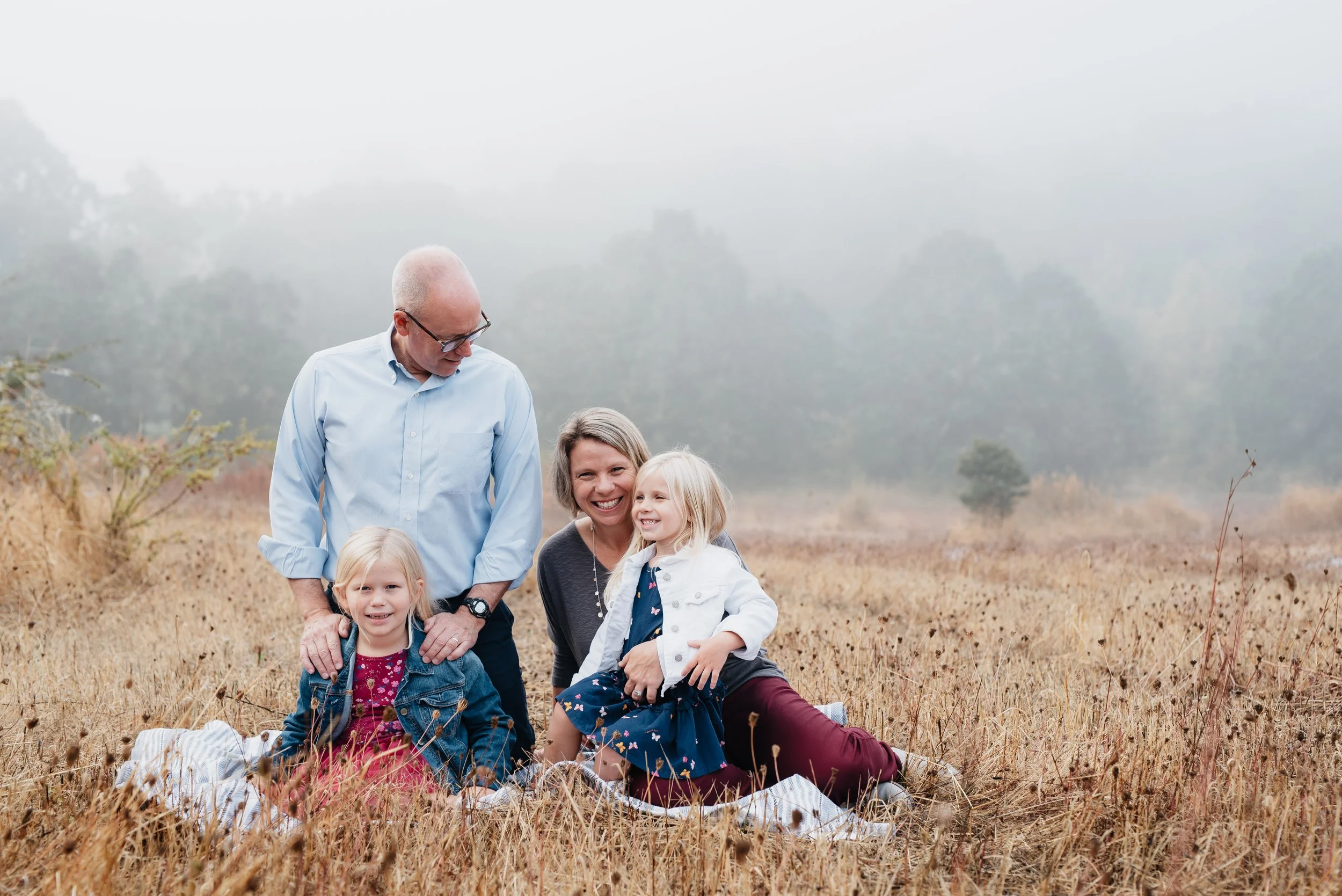 Family consisting of a father, mother, and two young girls sit on a blanket in an open field smiling