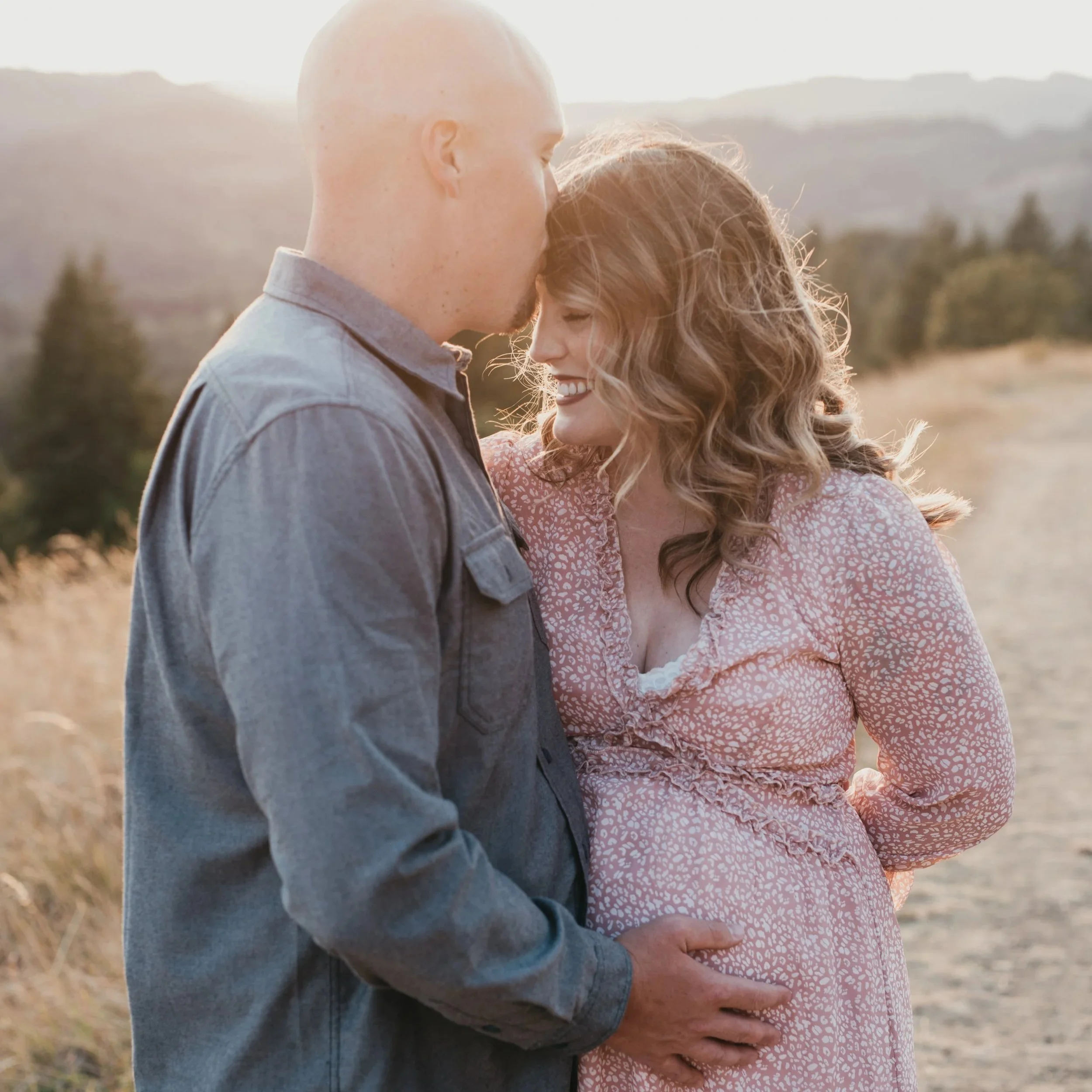 A husband kisses his pregnant wife on the forehead while the sun sets in the background.