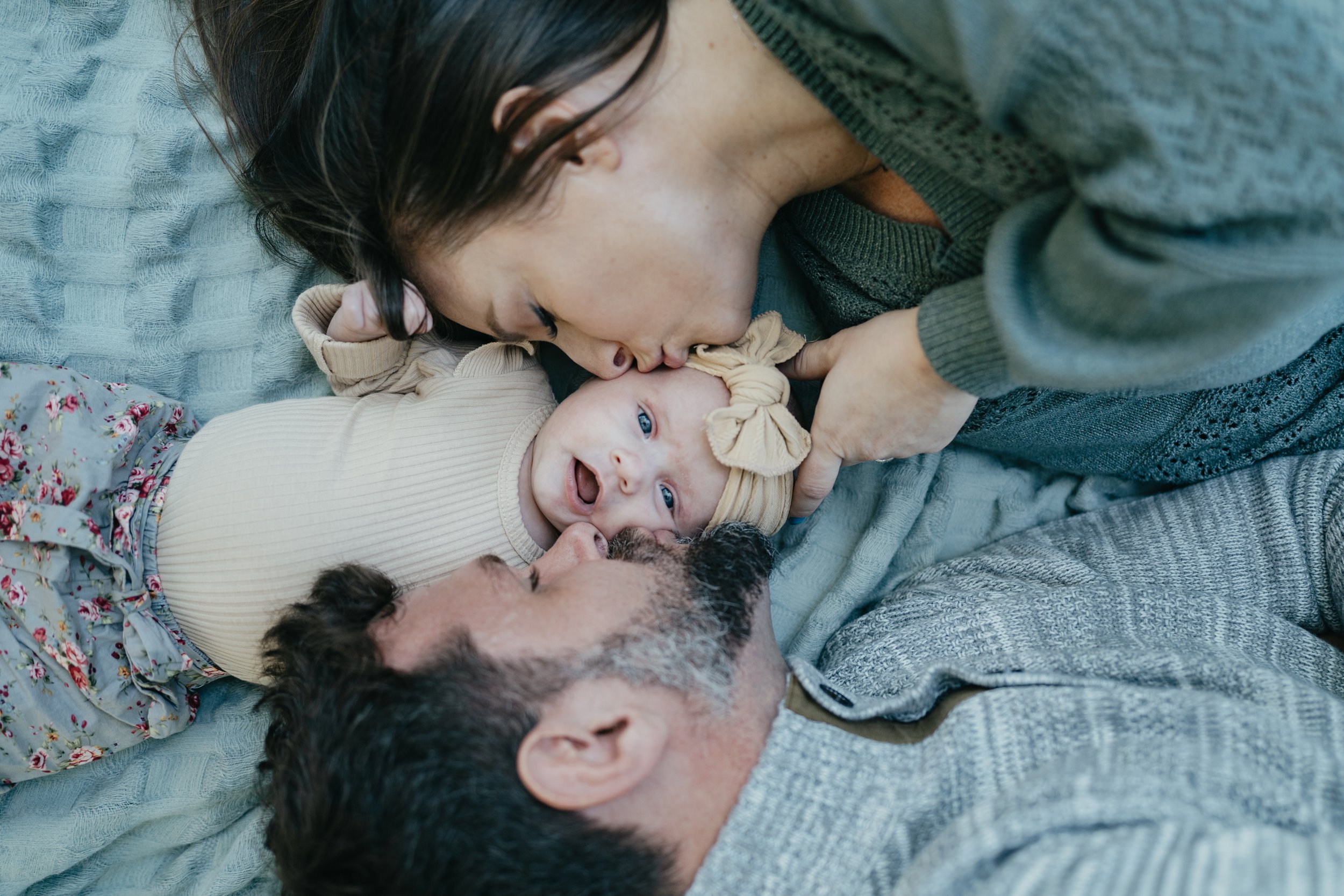 A mother and father are laying on a blanket kissing their one month old baby girl with big blue eyes on the cheek.