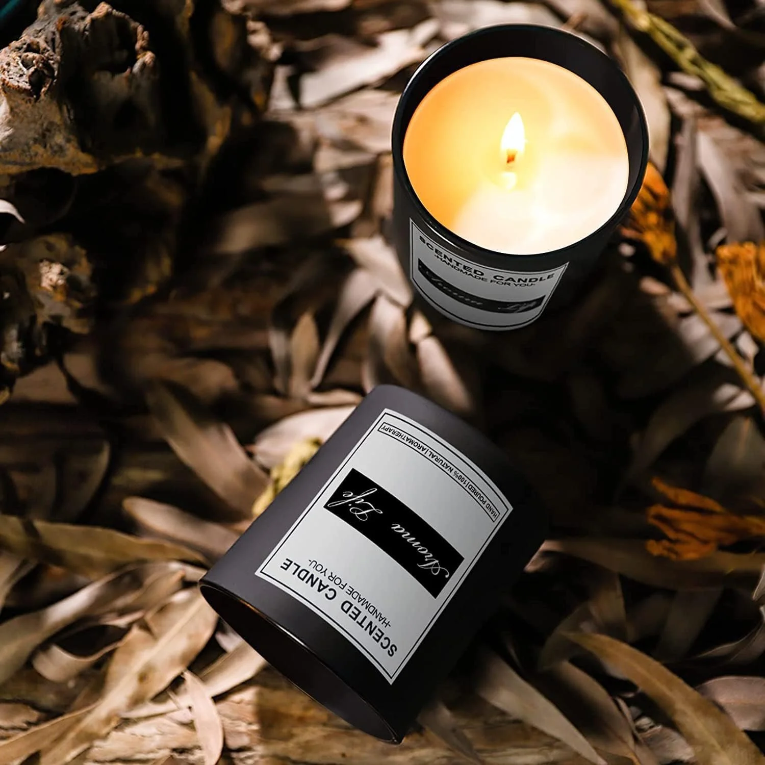 A lit scented candle labeled 'Scented Candle Handmade for You' and a black container labeled 'Scented Candle Handmade for You' placed on a bed of dried brown leaves.