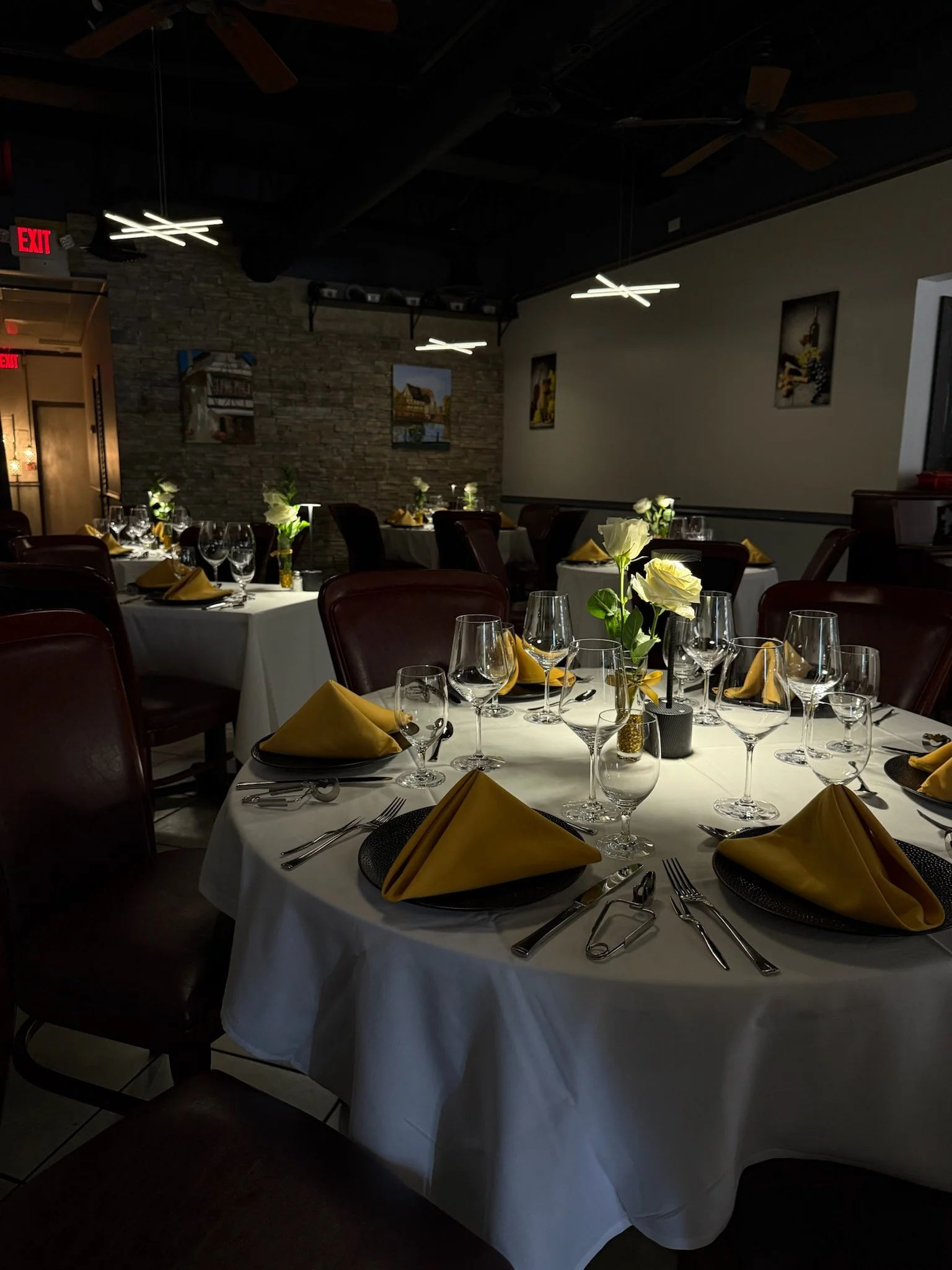 A dining area set for a formal event with tables covered in white tablecloths, yellow napkins, wine glasses, silverware, small floral centerpieces, and cushioned chairs.