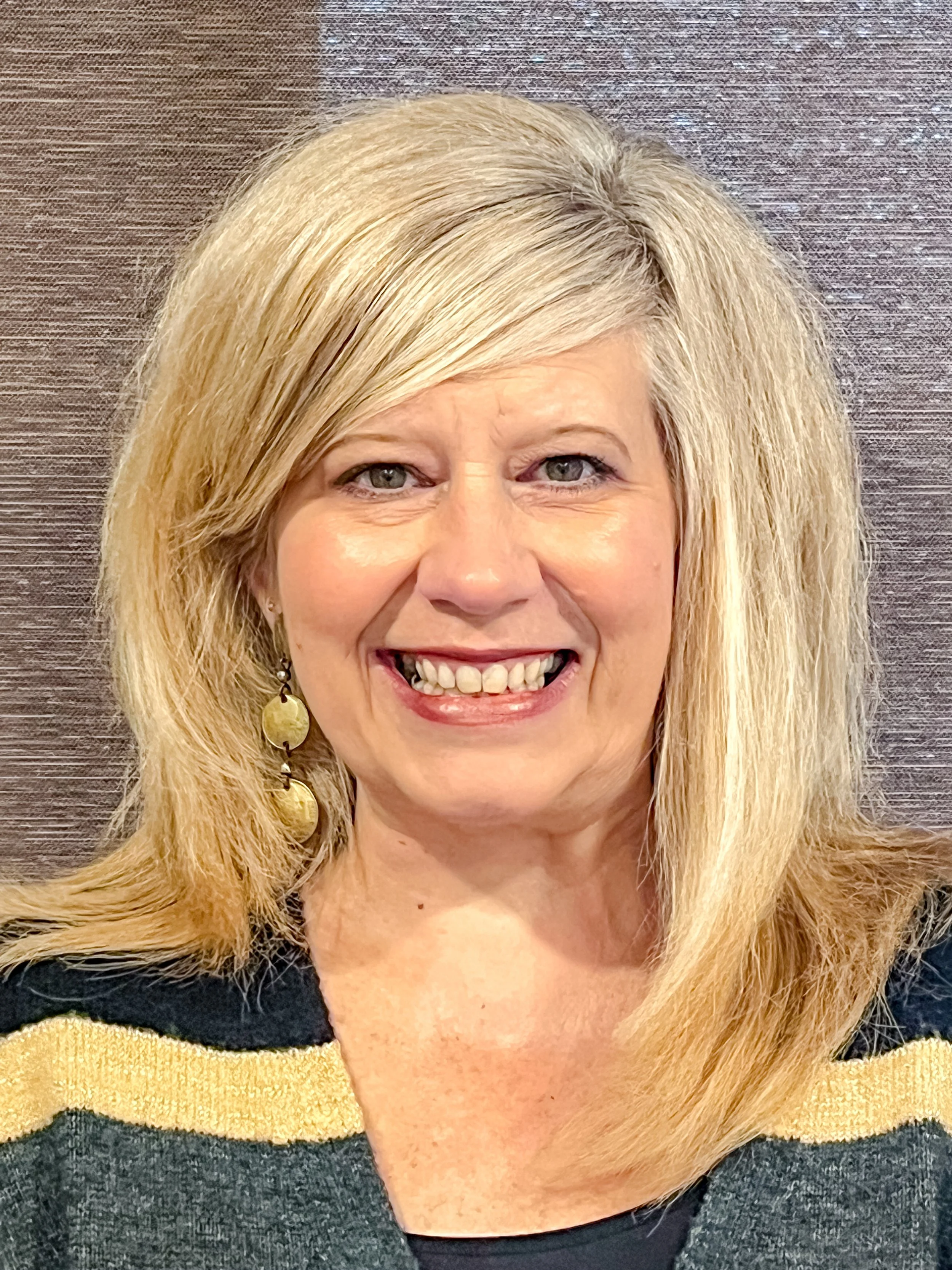 Close-up of smiling blonde woman with shoulder-length hair, wearing earrings, black and yellow top, against a textured background.