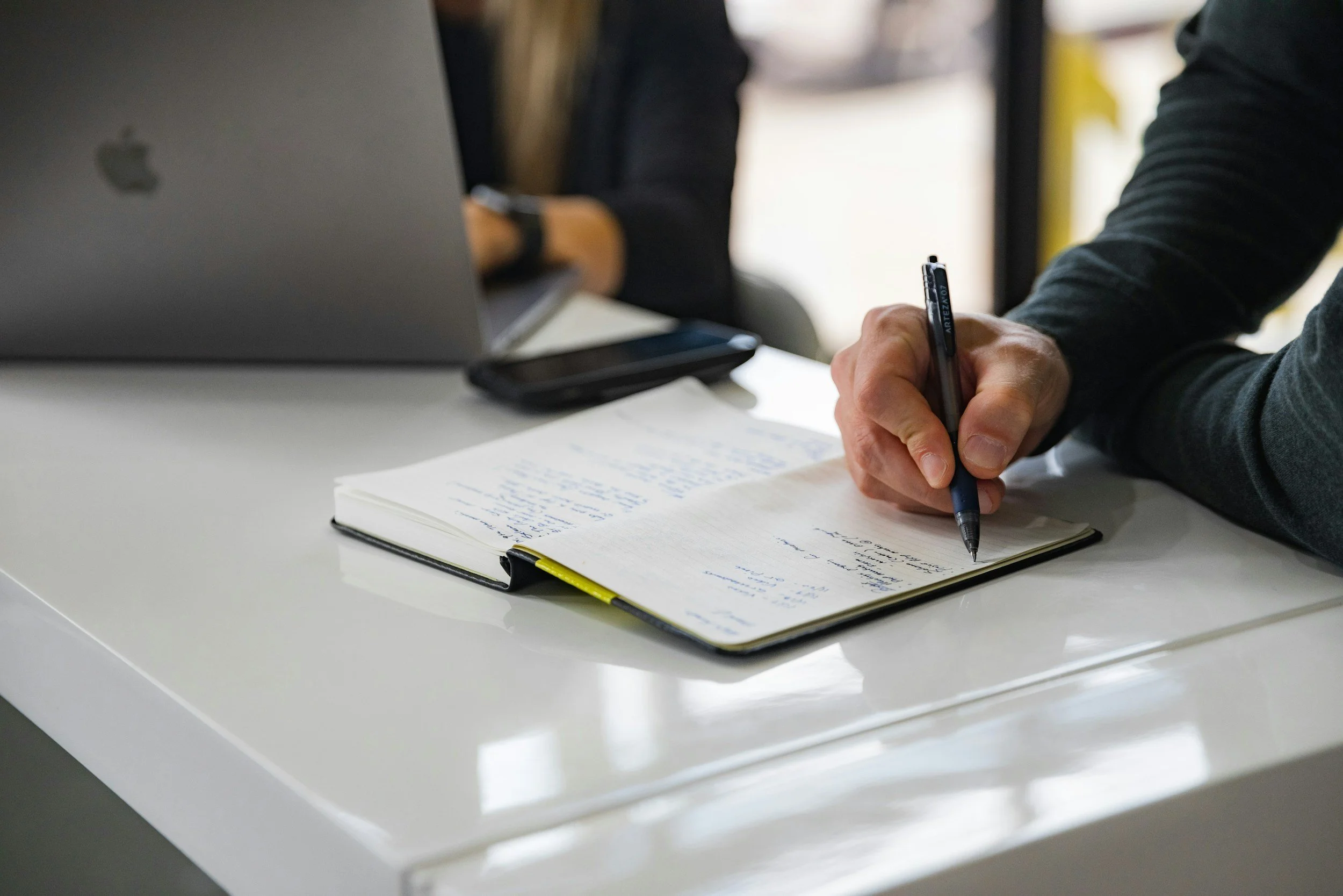 Person writing in a notebook with a black pen on a white table, with a laptop and a smartphone nearby.