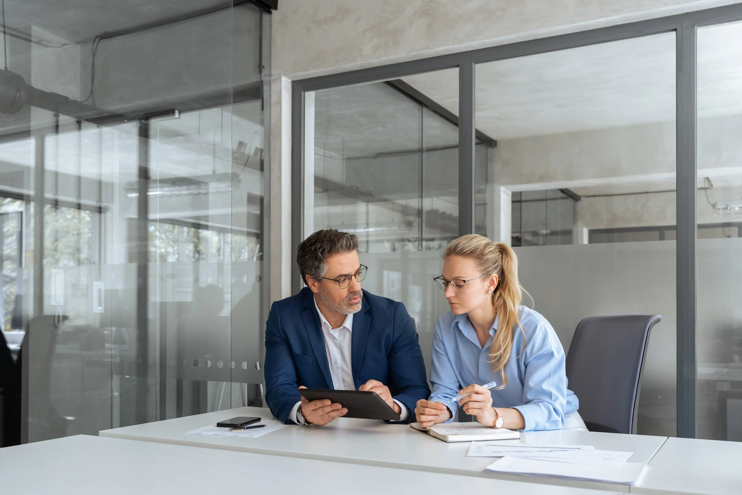 Two professionals, a man and a woman, sitting at a table in a modern office, engaged in a discussion while looking at a tablet and notes.