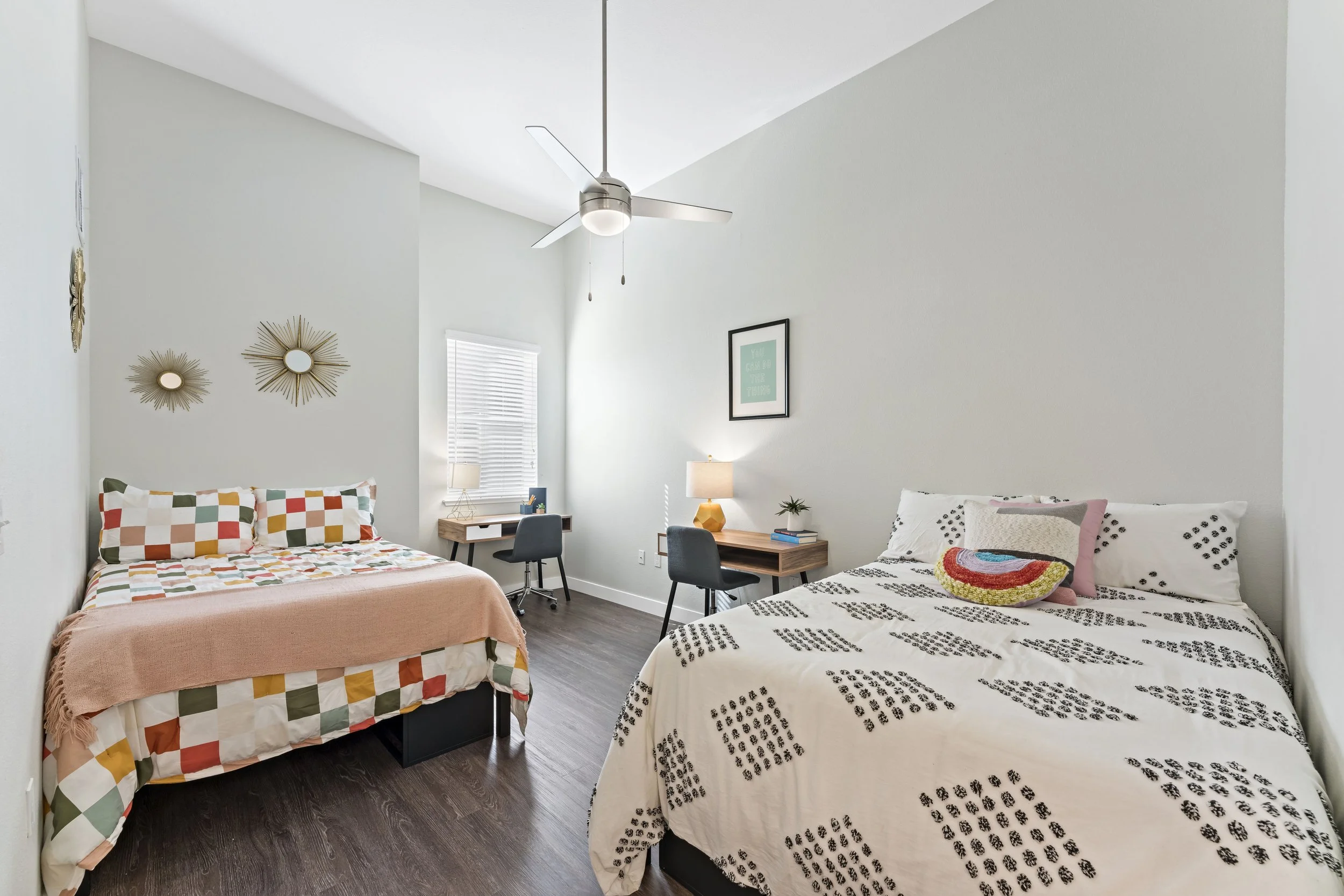 A bedroom with two beds, one with patchwork bedding and the other with black and white patterned bedding, a ceiling fan, two nightstands with lamps, and a small workspace with a desk, chair, and window.