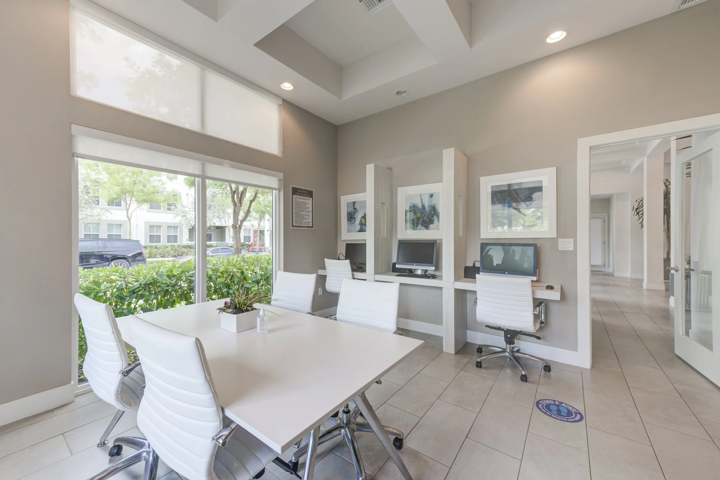 Modern office space with a white conference table, white chairs, large windows, computers, framed artwork, and indoor plants.