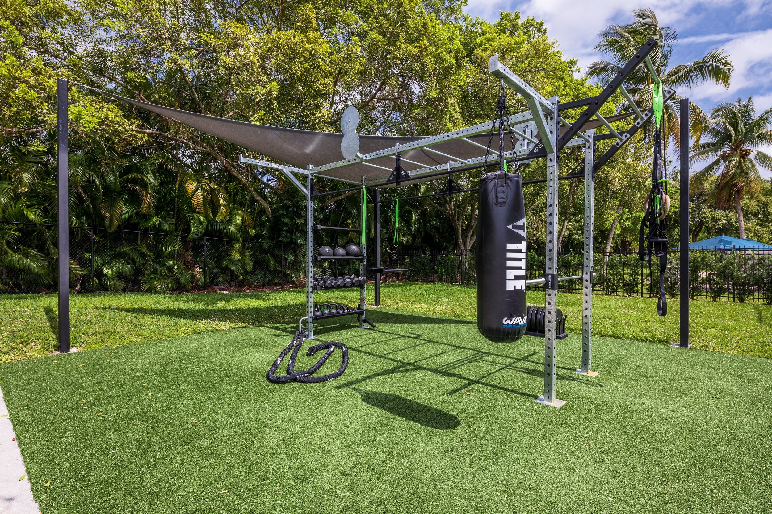 Outdoor fitness area with a metal frame structure, hanging punching bag, kettlebells, medicine balls, resistance bands, and a battle rope, surrounded by lush green trees and grass under a blue sky.