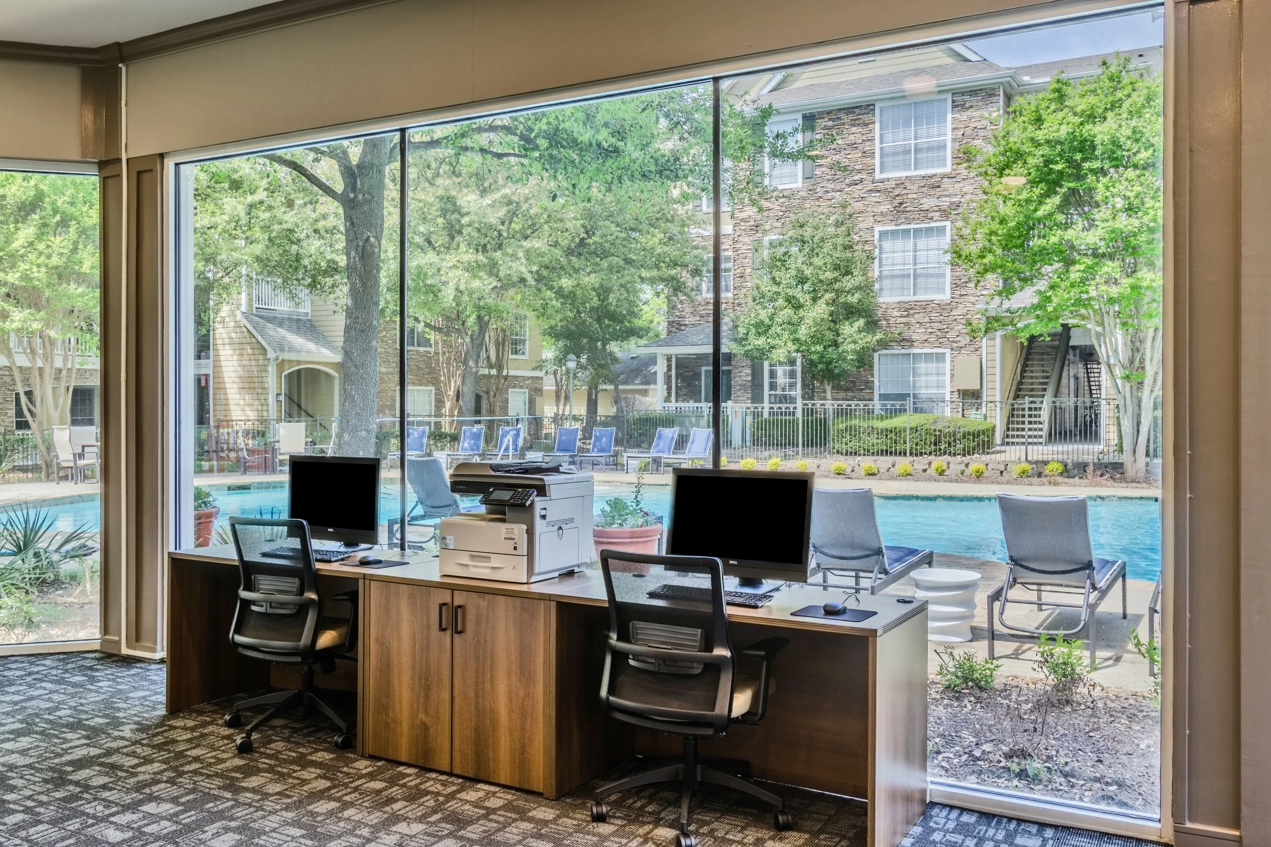 Office with two computers, a printer, and a view of a swimming pool outside through large glass windows.