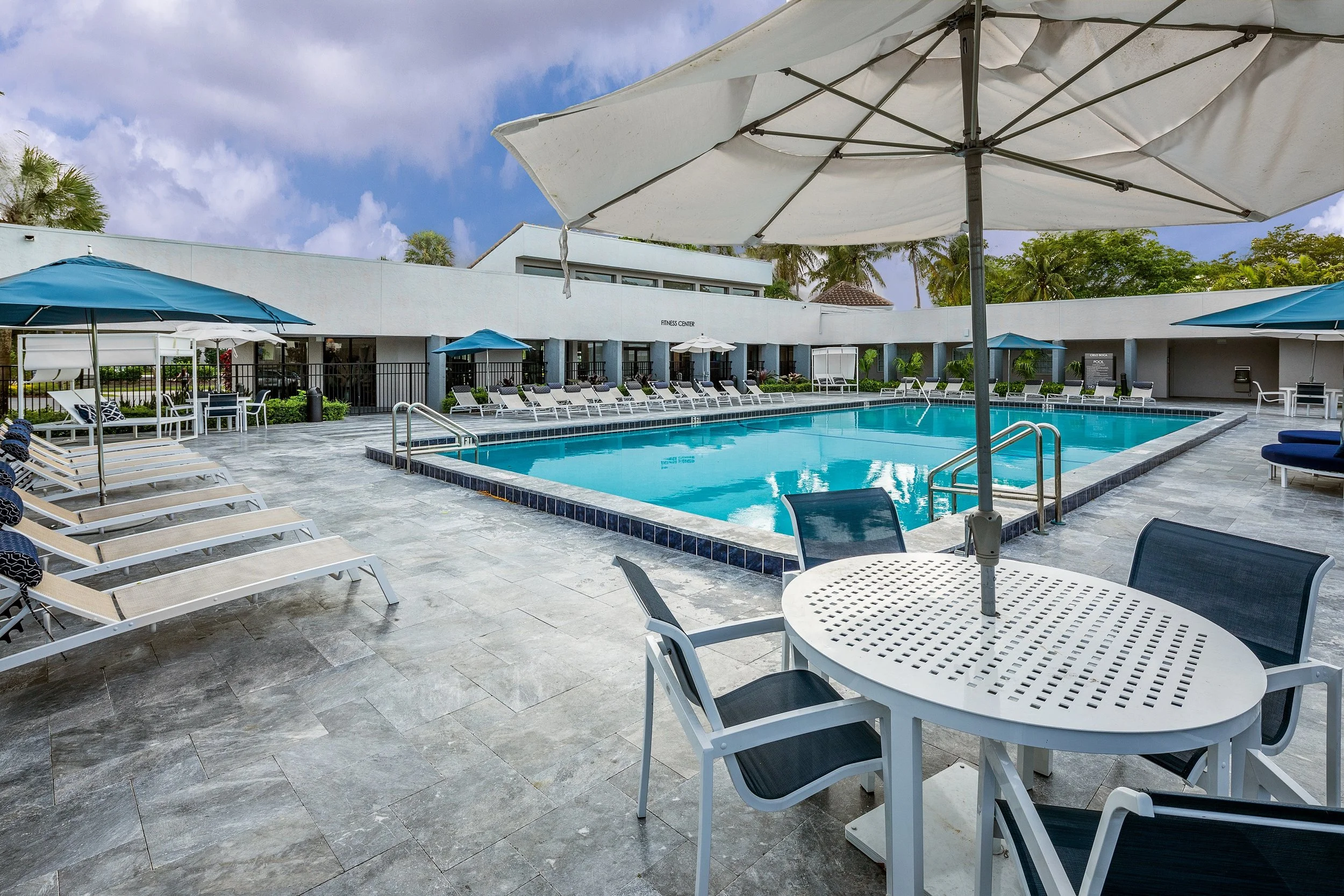 Empty outdoor swimming pool area with lounge chairs, umbrellas, and a table with chairs on a cloudy day