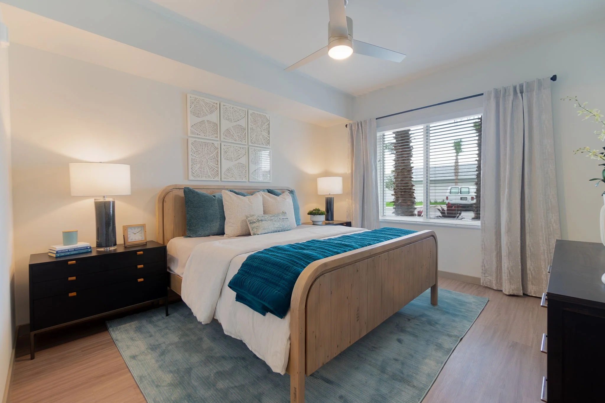 A bedroom with a wooden bed, white bedding, blue and white pillows, and a blue throw blanket. Two black nightstands with lamps, a window with blinds and curtains, and a ceiling fan.