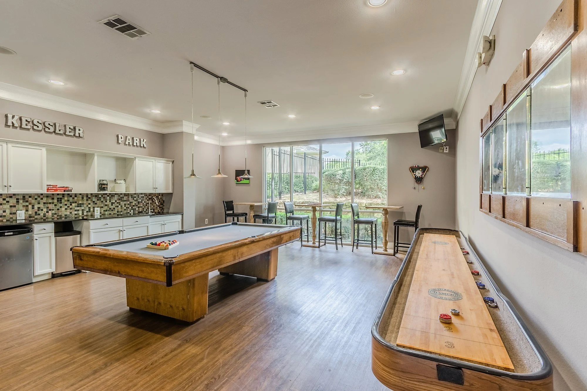 Indoor recreation room with a pool table, air hockey table, seating area near large window, and kitchen area with white cabinets and a tile backsplash.