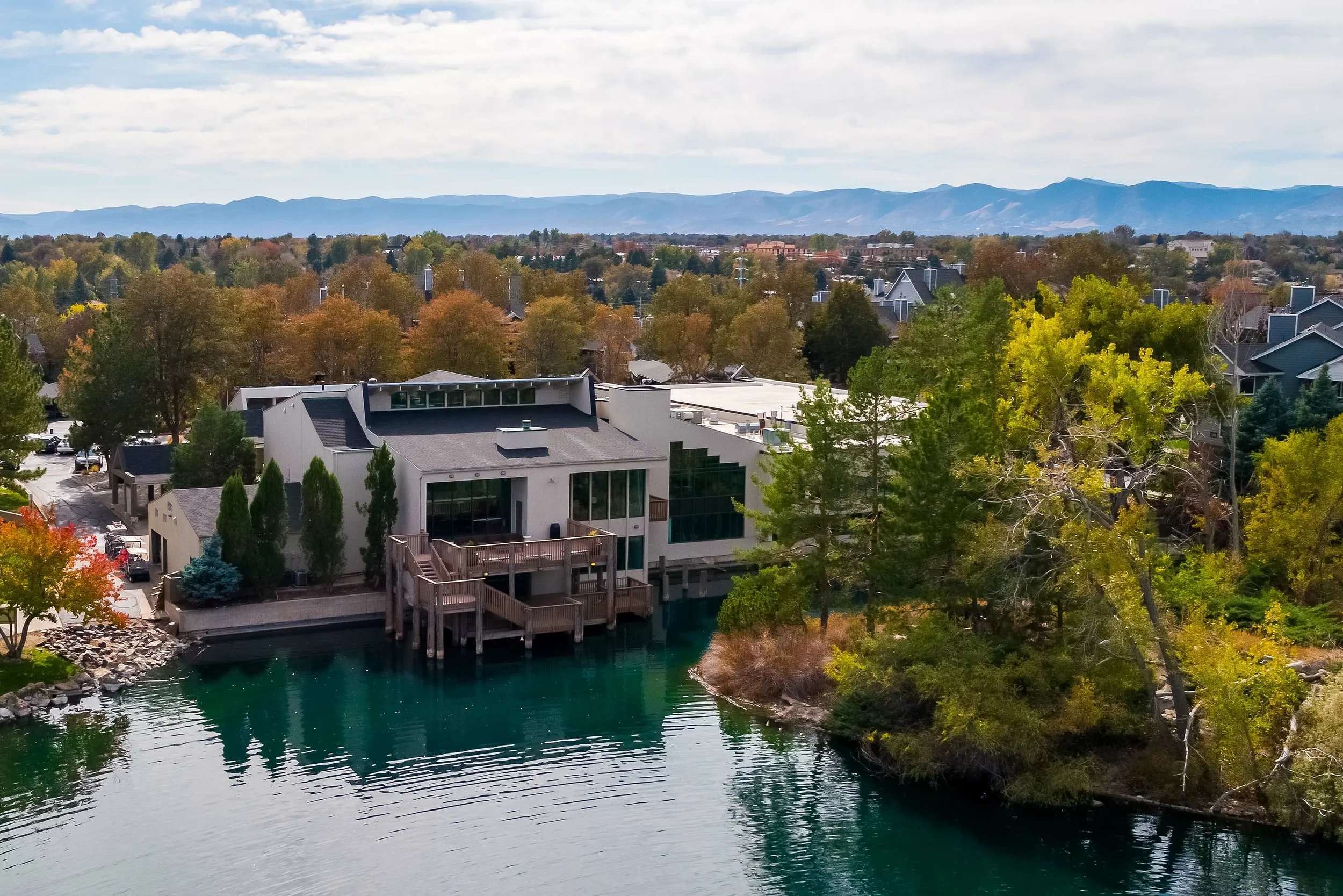 Aerial view of a modern house with large glass windows situated by a small lake, surrounded by autumn-colored trees and residential neighborhood, with mountains in the distance under a partly cloudy sky.