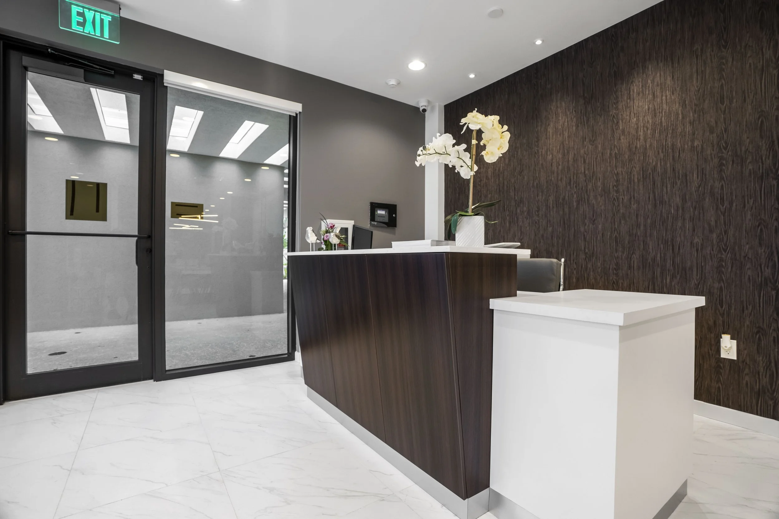 Reception area with a dark wooden counter, a white side table with a potted white orchid, gray and dark wood walls, and an exit sign above glass doors.