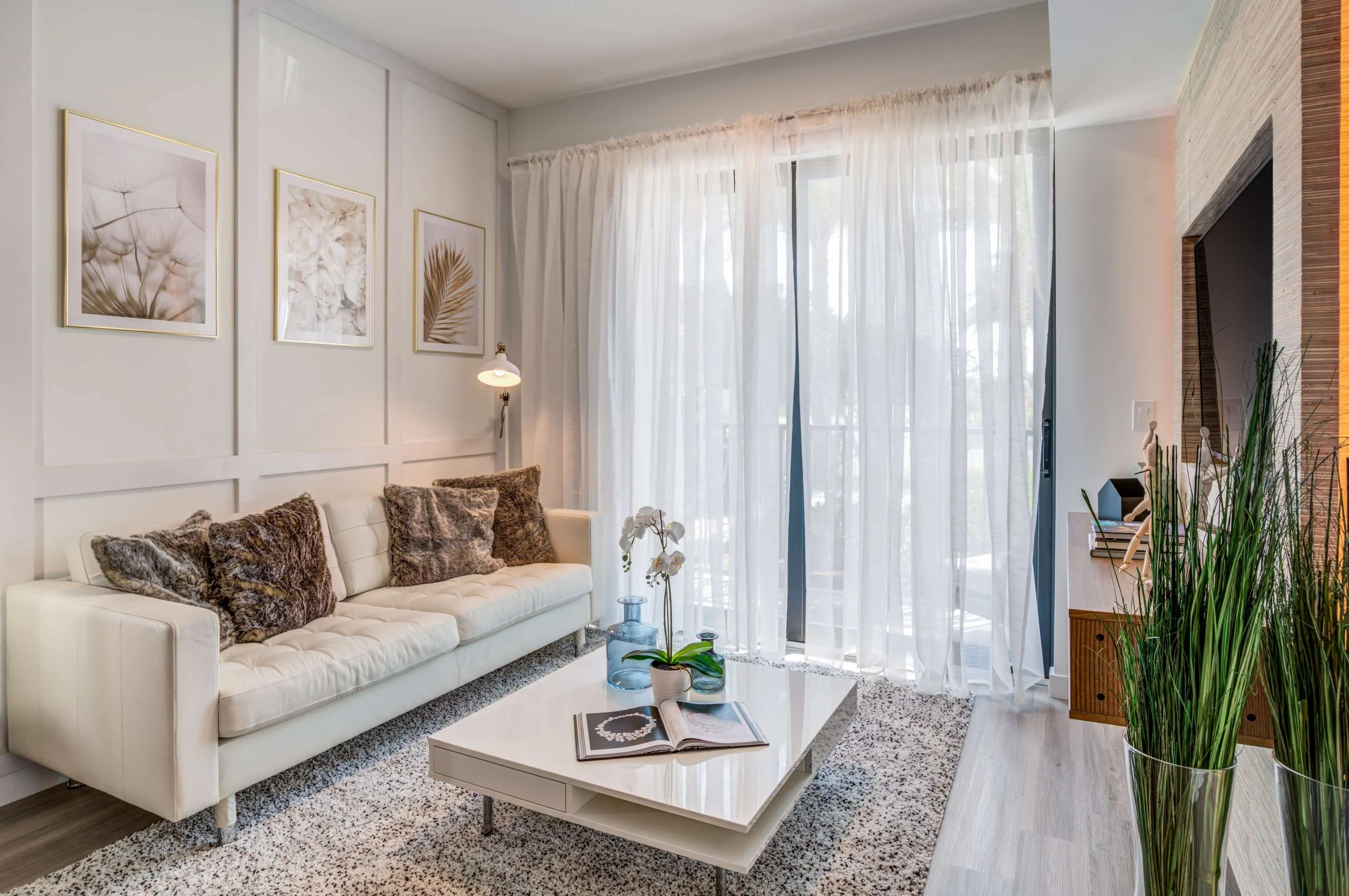 Modern living room with white sofa, fur pillows, glass coffee table with magazines and a potted orchid, large windows with sheer curtains, framed wall art, green plants, and a flat-screen TV.