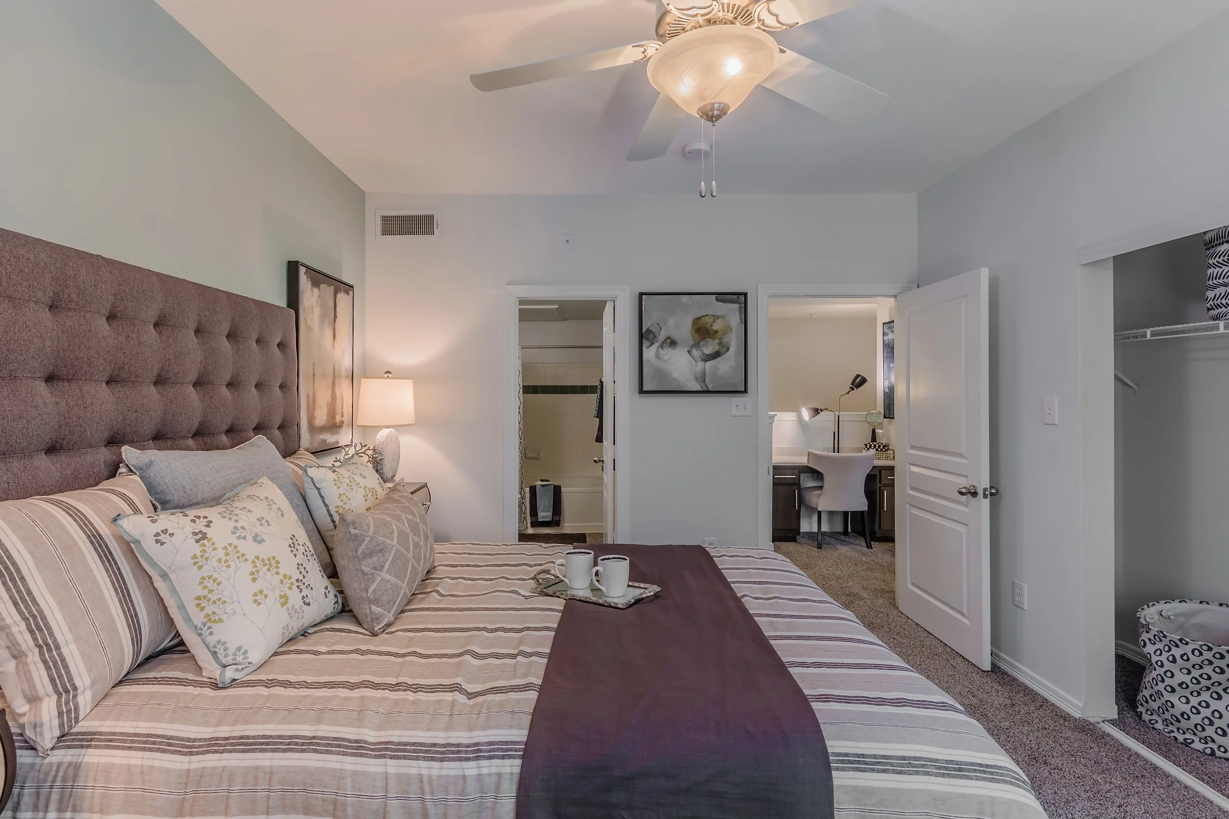 A bedroom with a large bed, multiple pillows, a bedside lamp, a ceiling fan, a small desk with a chair and desk lamp, an open closet, and a bathroom in the background.