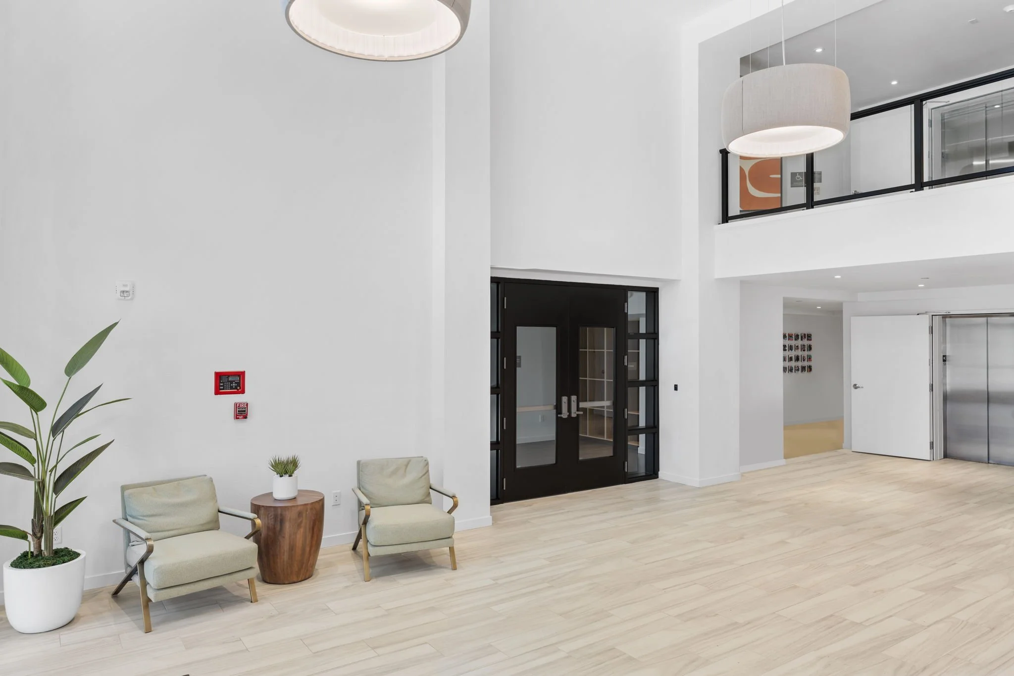 Modern interior with light wood flooring, white walls, large black double door with glass panels, wall-mounted elevator, green potted plant, two beige chairs with a small wooden table and plant, white open door, and circular hanging lights.