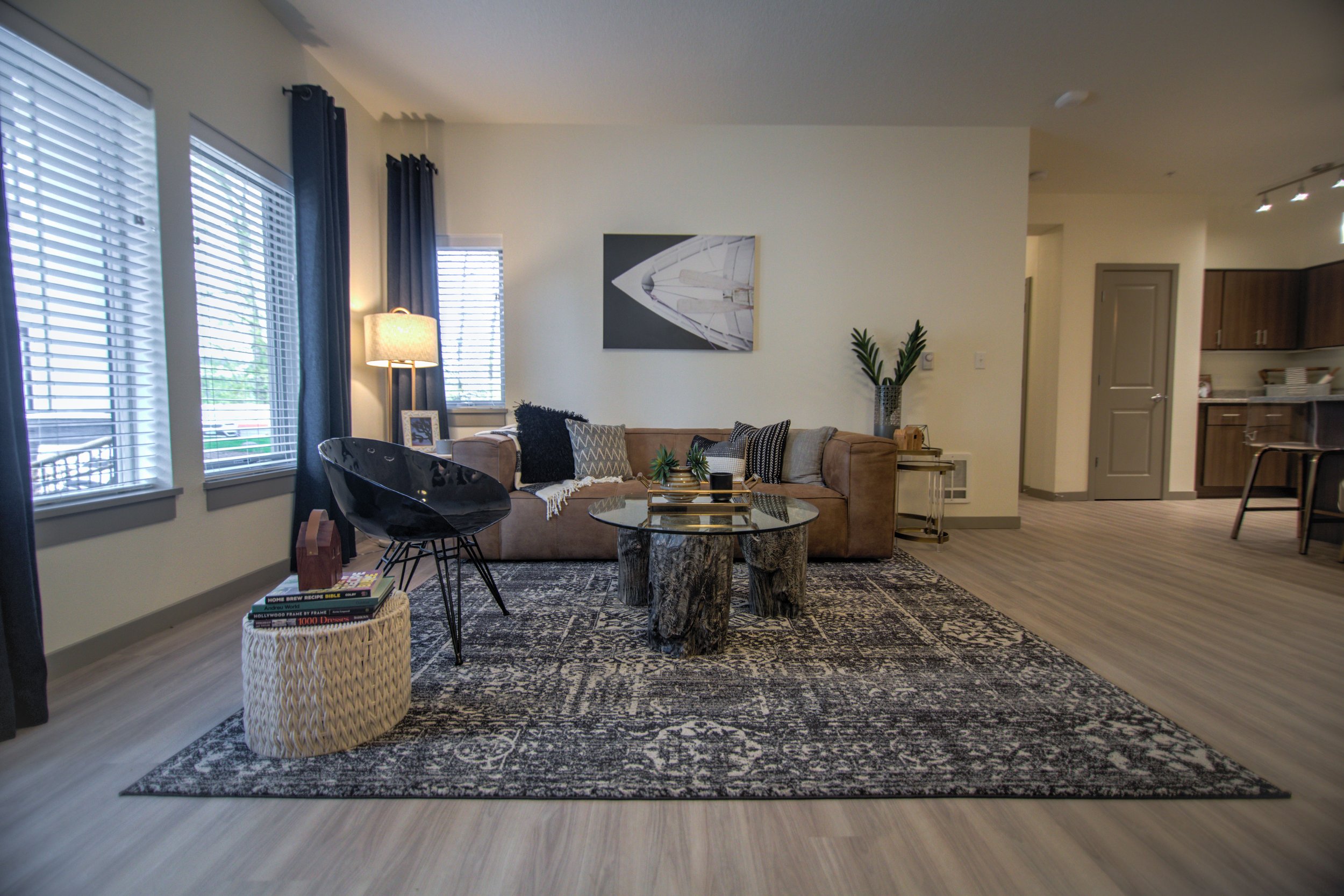 Living room with beige sofa, black armchair, glass coffee table, patterned area rug, and large windows with blinds and dark curtains.