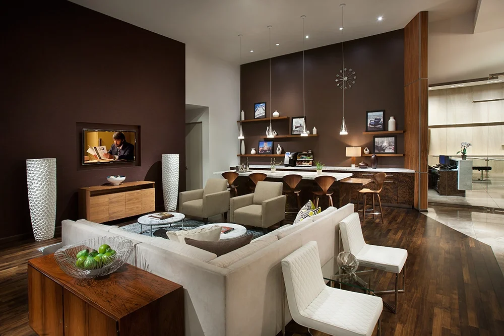 Modern living room with beige and white furniture, dark purple accent wall, wall-mounted TV, wooden shelving, and a kitchen area with pendant lighting and bar stools.