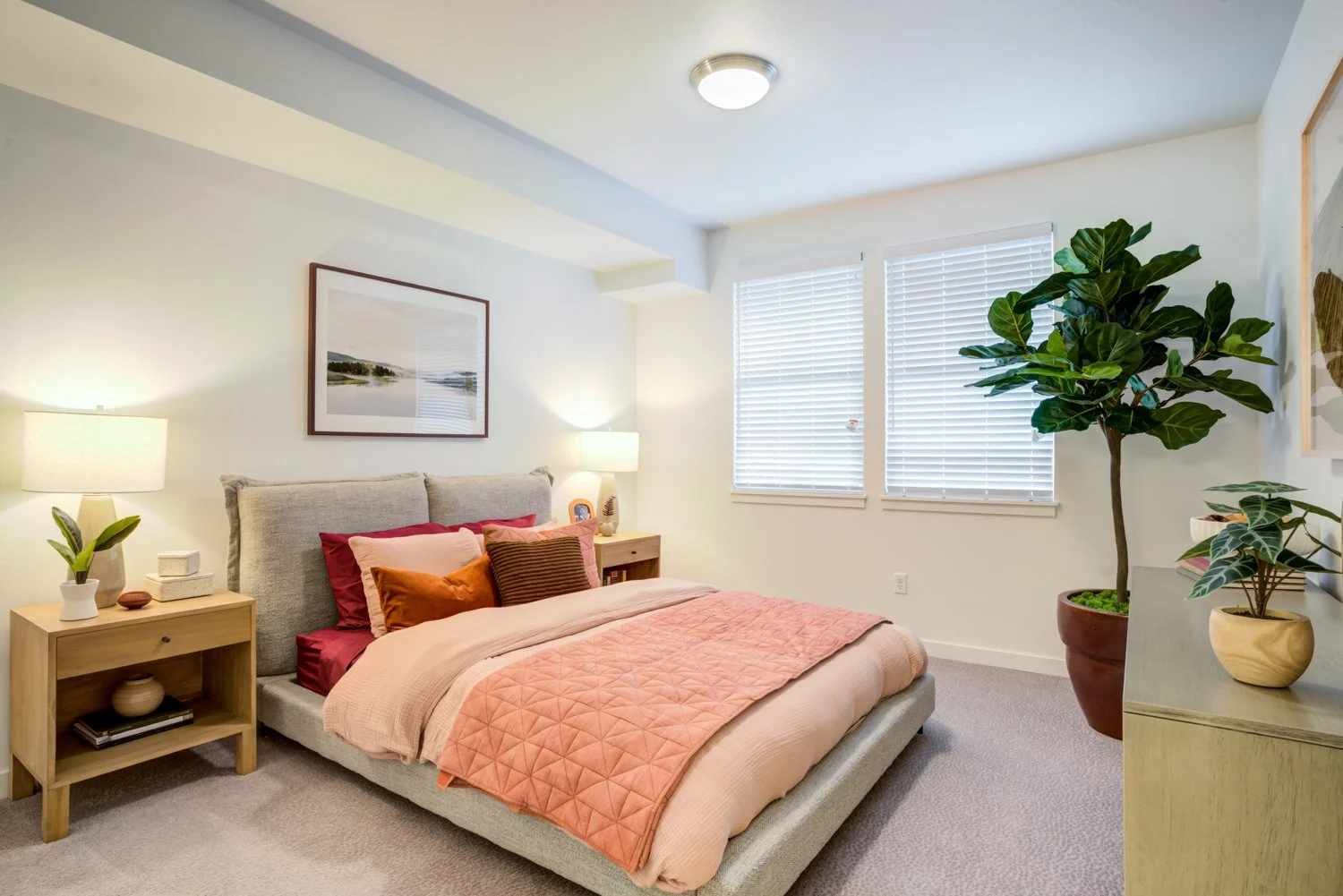 A modern bedroom with a bed, bedside tables with lamps, a large potted plant, and a framed landscape picture on the wall.