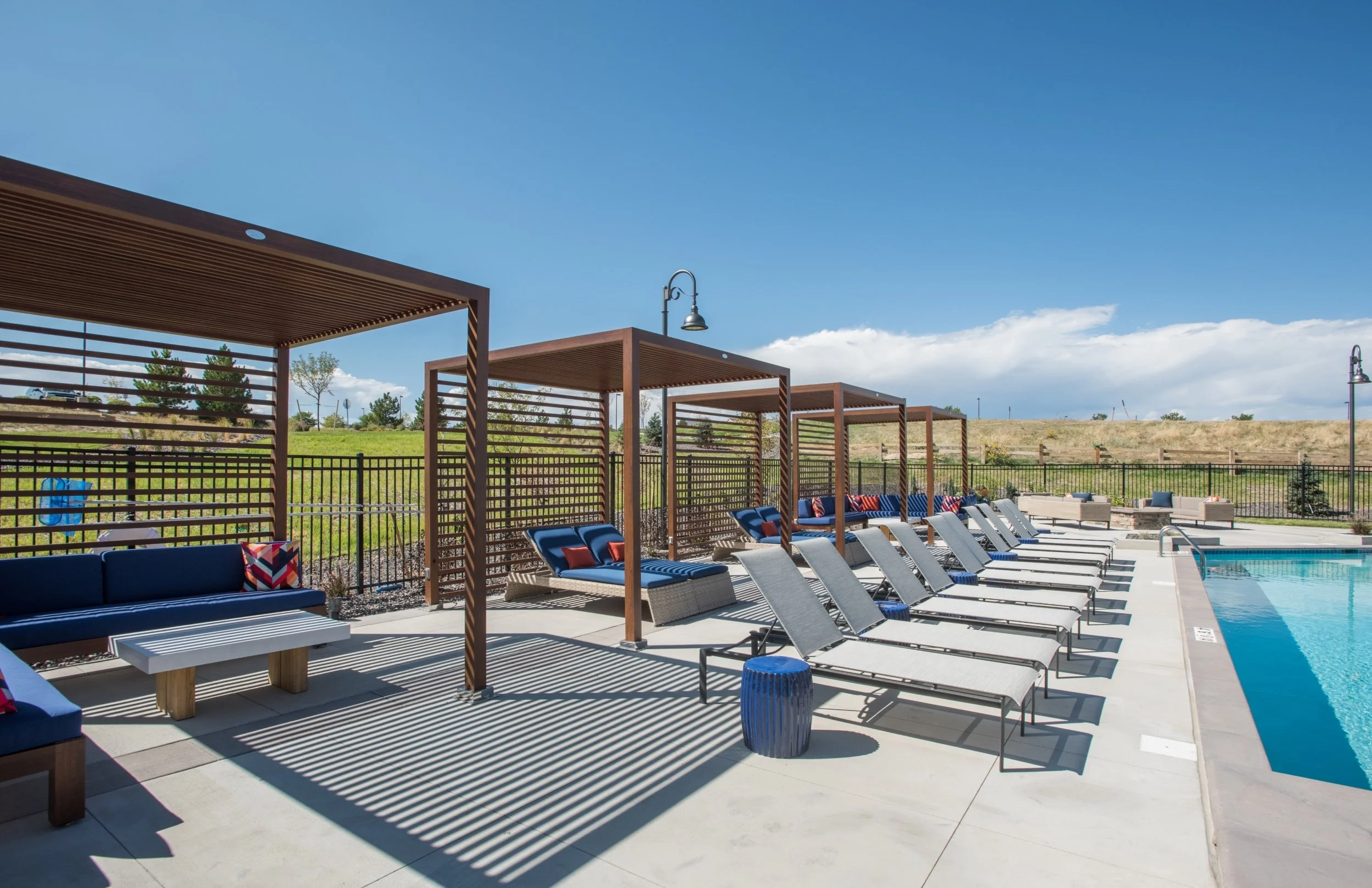 Swimming pool area with lounge chairs and shaded cabanas under a bright blue sky.