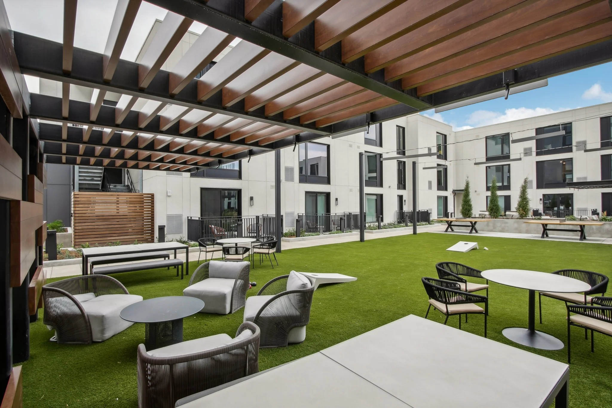 Outdoor patio with green artificial grass, black metal tables and chairs, sofas, and wooden pergola, adjacent to modern white apartment buildings.