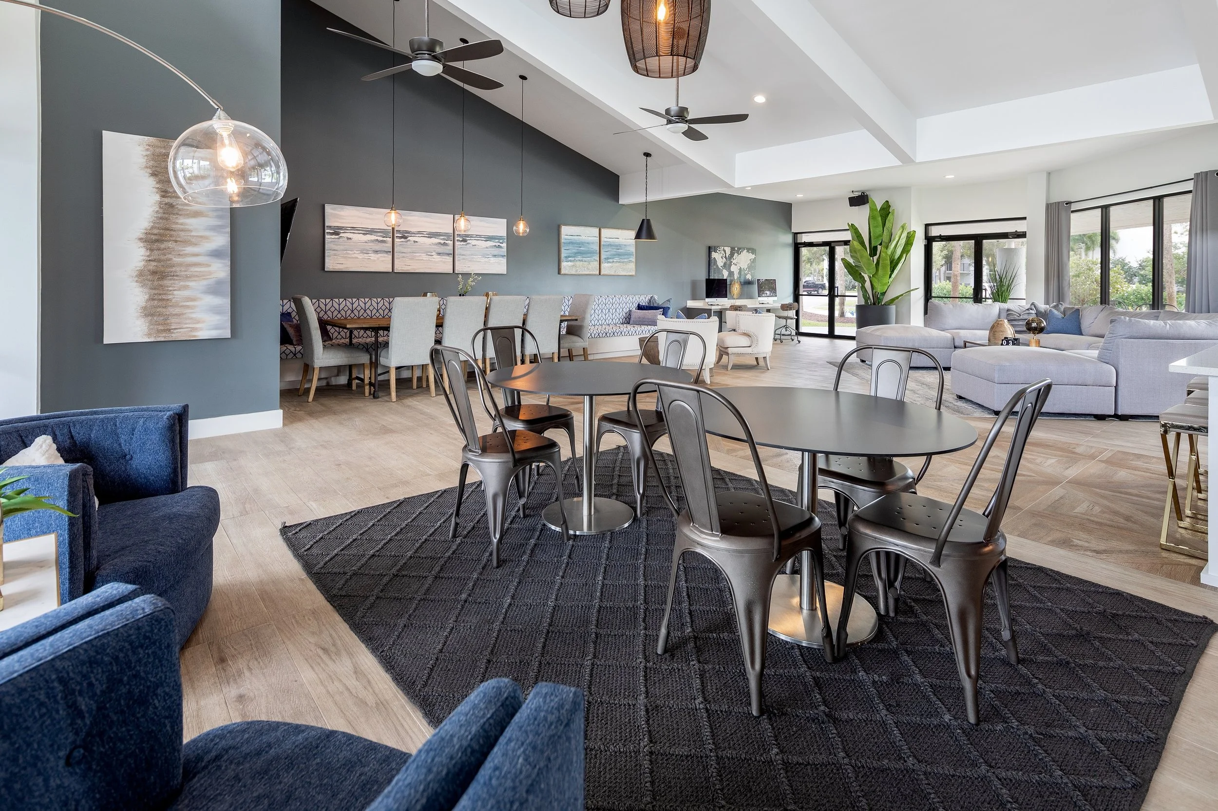 Modern, open-concept living room and dining area with tables, chairs, sofas, large windows, ceiling fans, and decorative lighting.